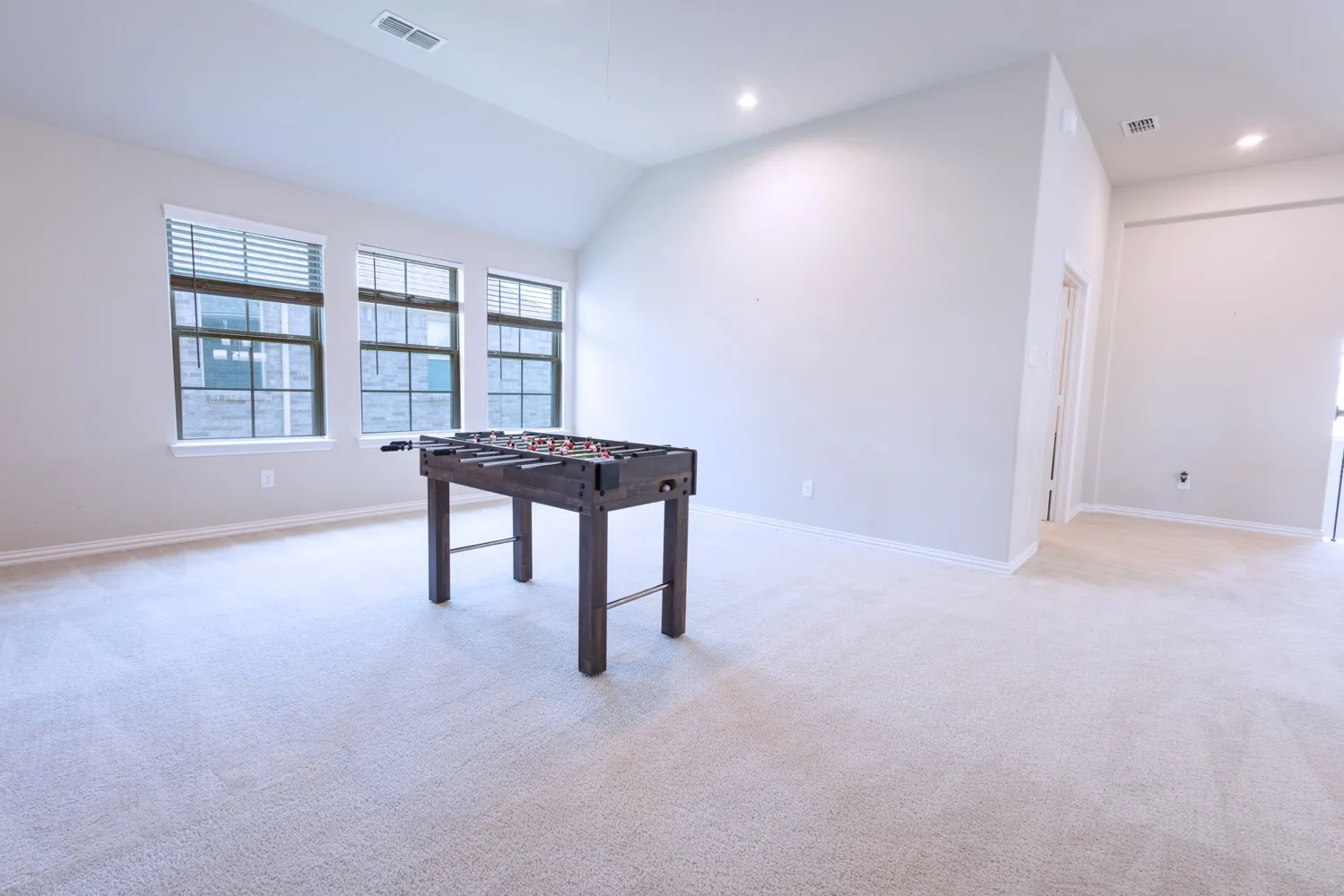 Spacious room featuring vaulted ceilings, light-toned carpeting, recessed lighting, and a trio of windows with blinds