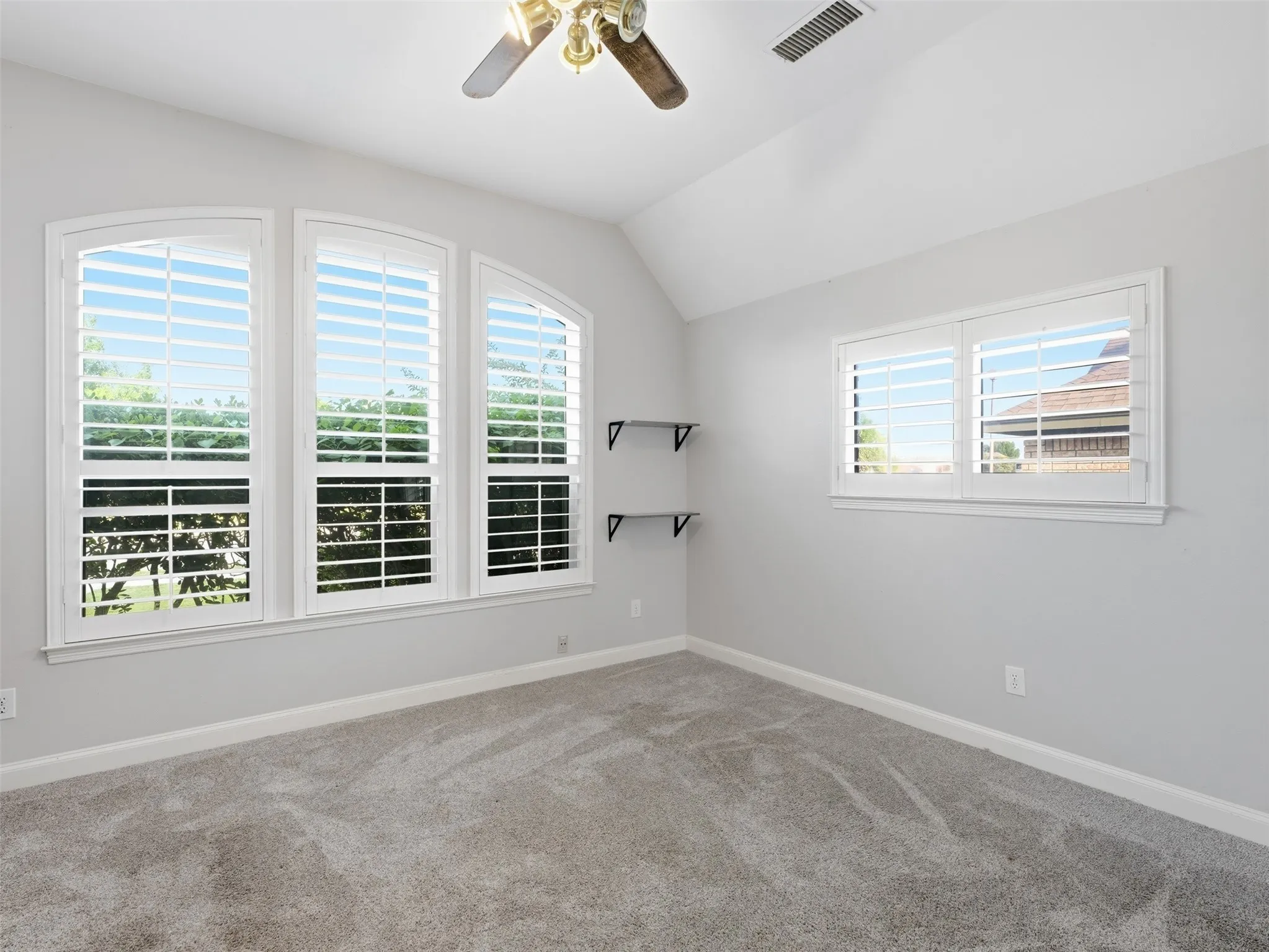 Room featuring arched windows with white plantation shutters, vaulted ceiling, ceiling fan, and grey carpet