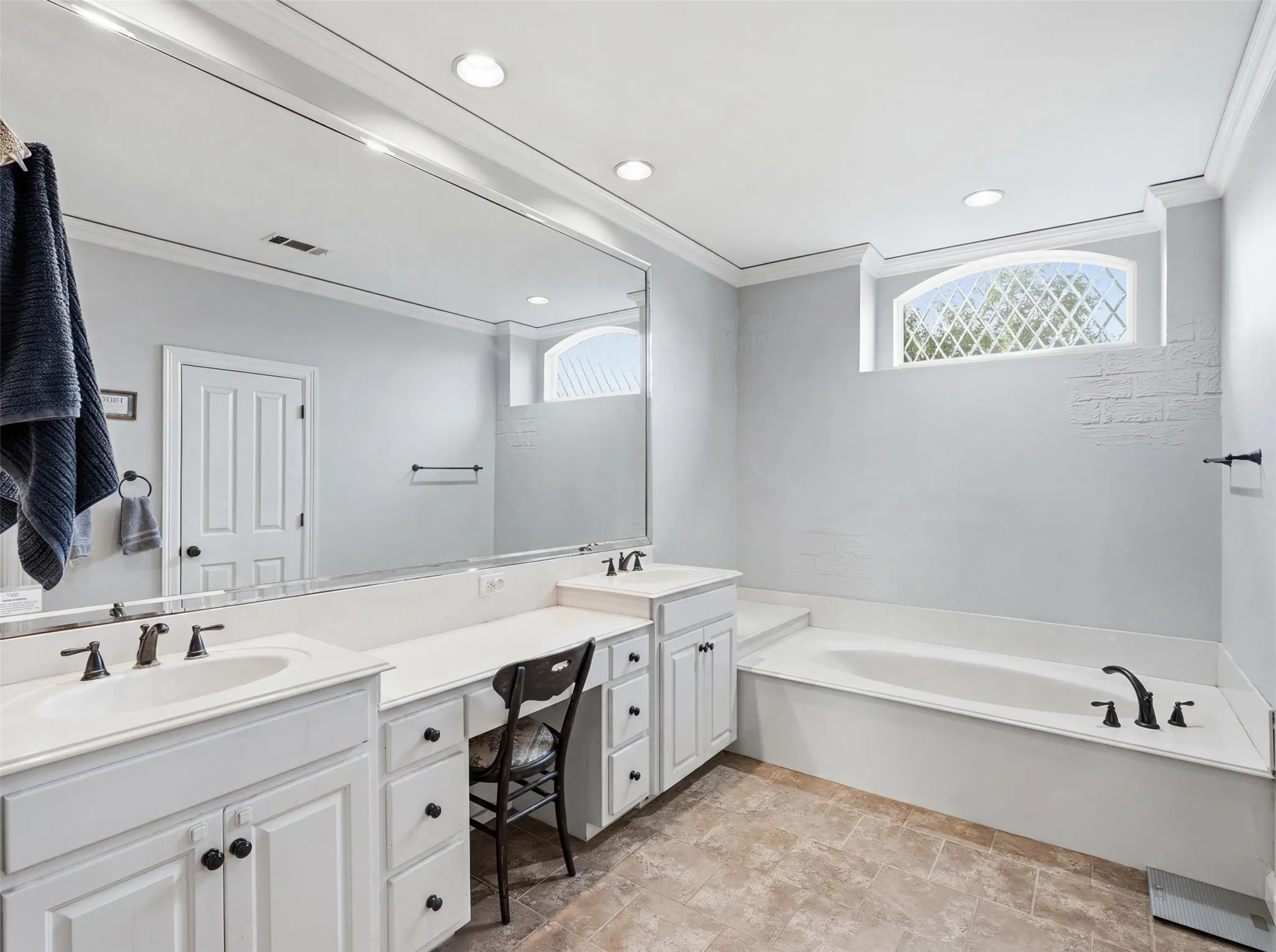 Spacious primary bathroom featuring a built-in tub, dual vanities with white cabinetry, extensive counter space, and a decorative arched window