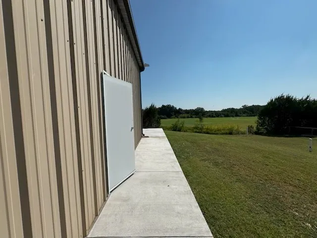 Tan corrugated metal siding featuring a white entry door, concrete walkway, and expansive green lawn