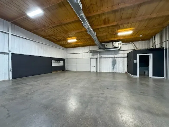 Spacious open warehouse area featuring a polished concrete floor, wood-paneled ceiling, exposed ductwork, fluorescent lighting, and an enclosed restroom facility