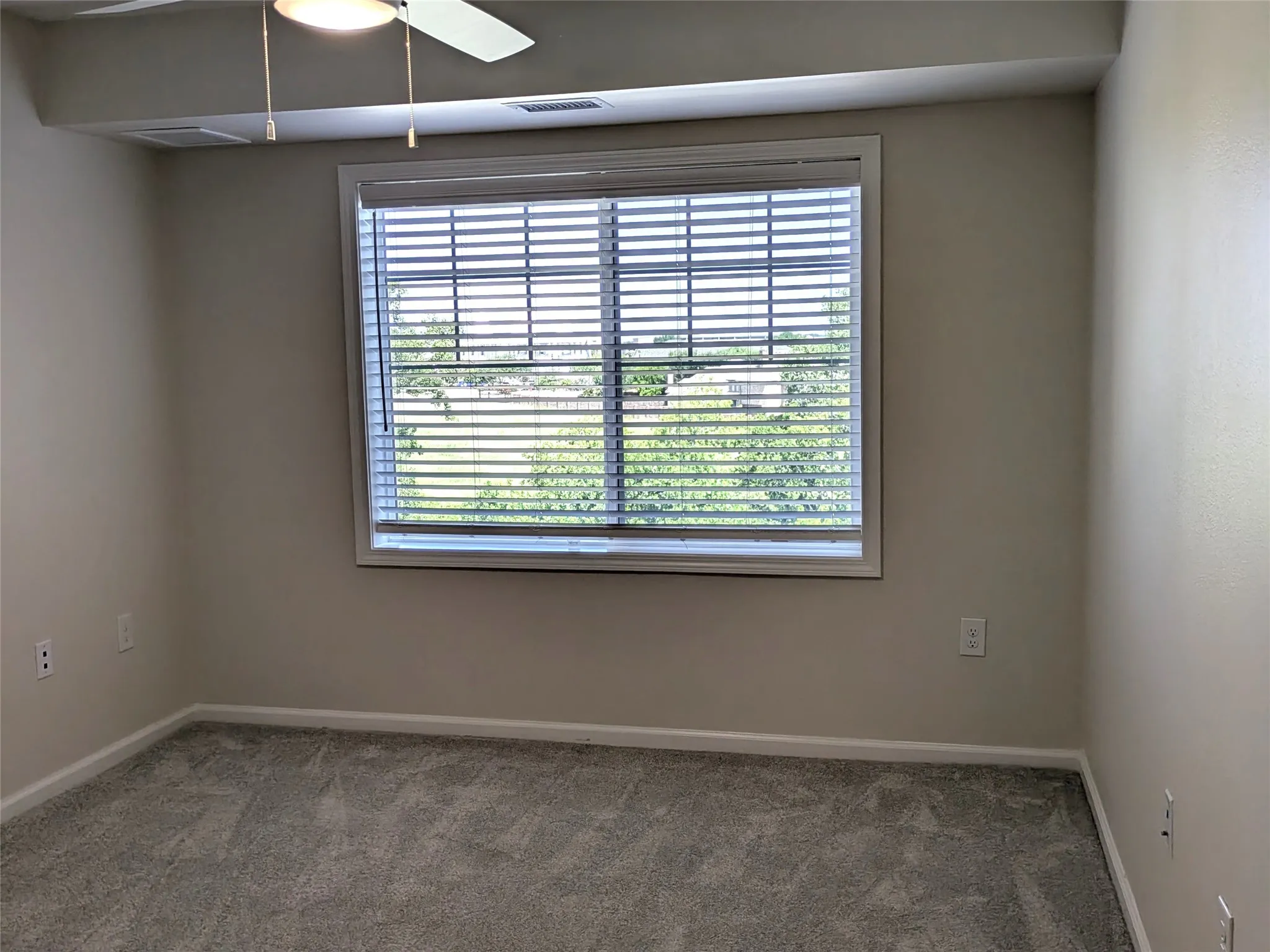 Neutral-toned room featuring a window with horizontal blinds, light gray textured carpet, white baseboards, and a ceiling fan with integrated lighting