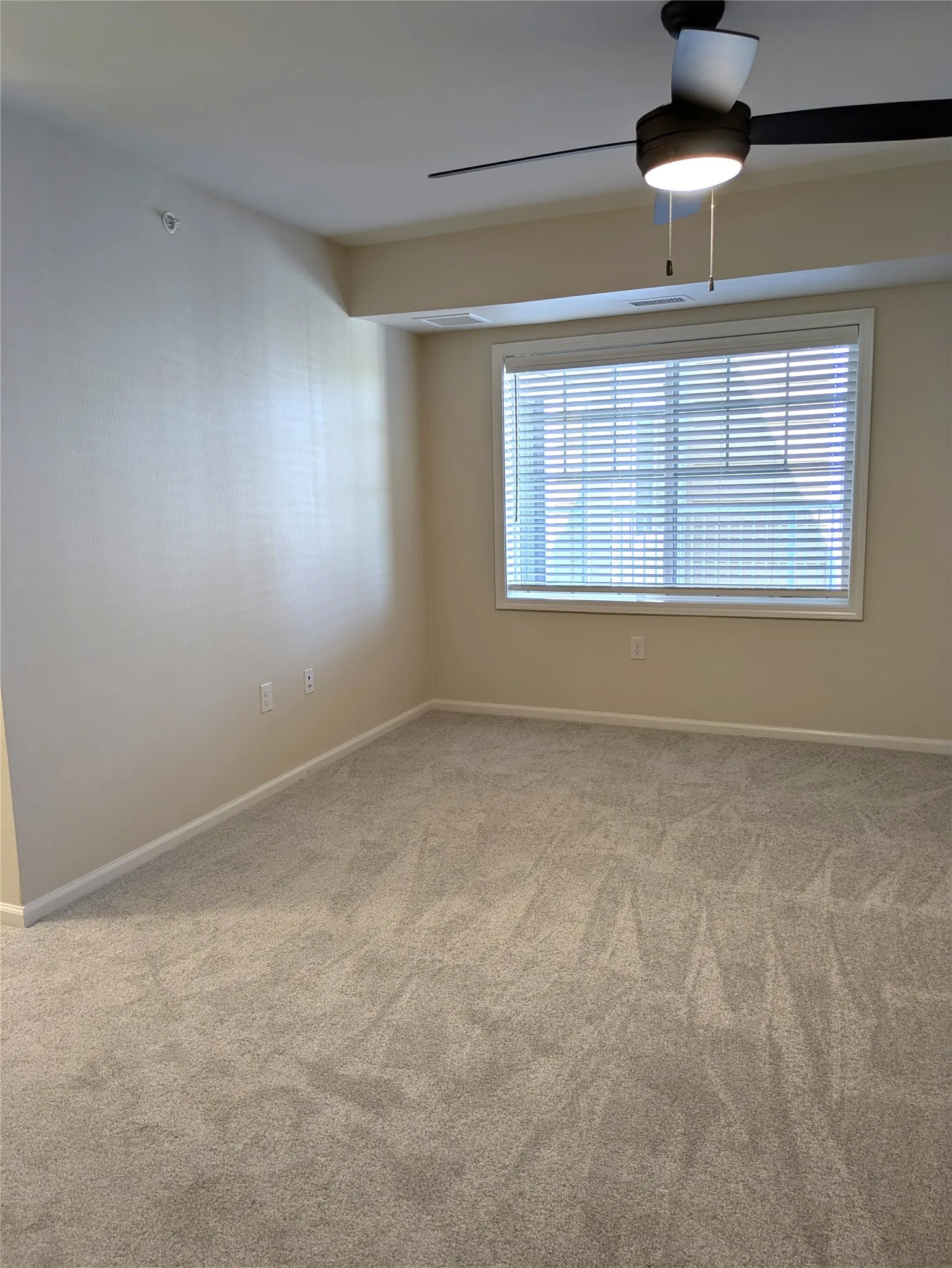 Neutral-toned room featuring a large window with horizontal blinds, overhead ceiling fan with lighting, and light grey patterned carpeting