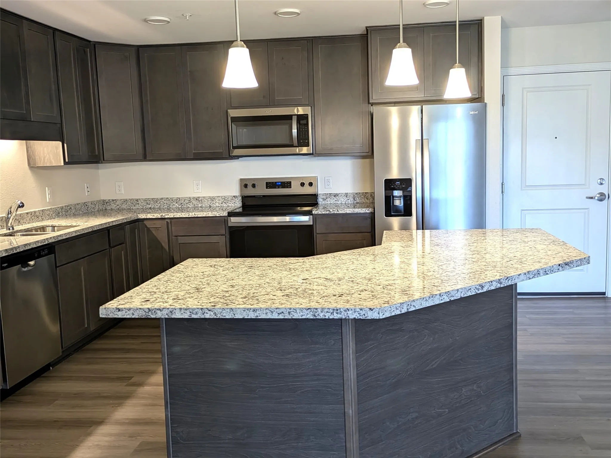 Kitchen featuring dark wood-finish cabinetry, light-toned countertops, stainless steel appliances, a center island with pendant lighting, and wood-finish flooring