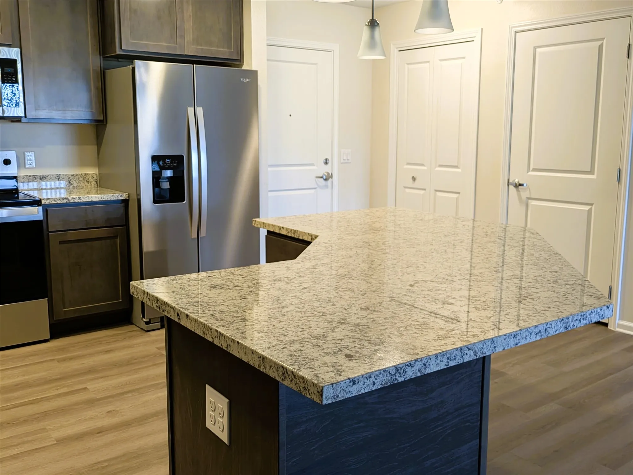 Kitchen island featuring a light-toned speckled countertop, dark wood-finish cabinetry, stainless steel side-by-side refrigerator, wood-finish flooring, and recessed ceiling lighting