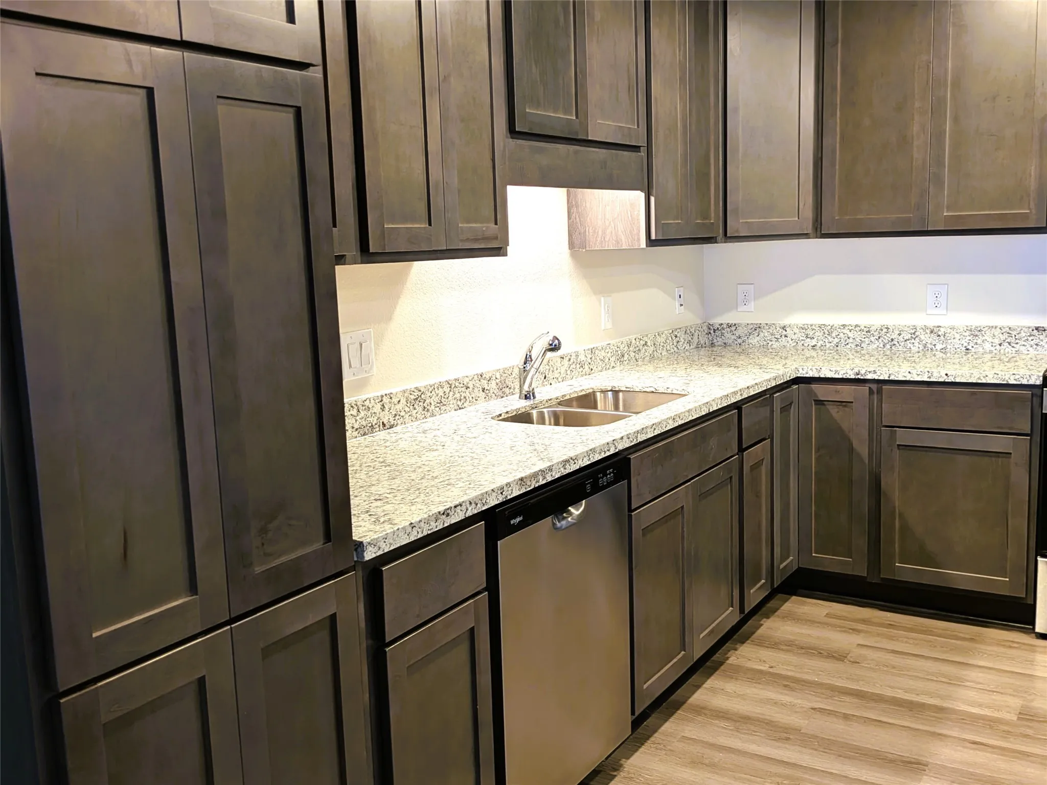 Kitchen featuring wood-finish shaker-style cabinetry, light-toned speckled countertops, a stainless steel double basin sink with a chrome faucet, and a stainless steel dishwasher
