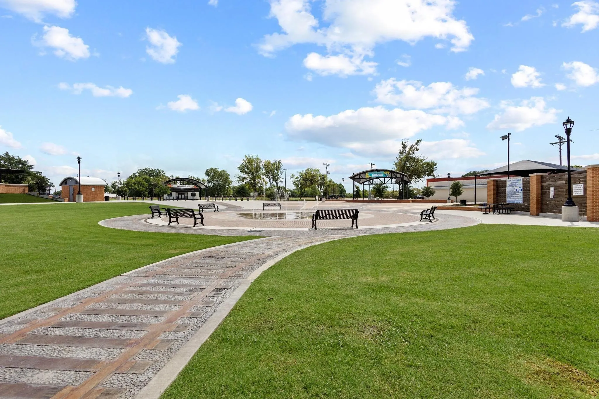 Expansive green space featuring a curved paved pathway, a central plaza with a water feature, multiple seating benches, and decorative street lamps