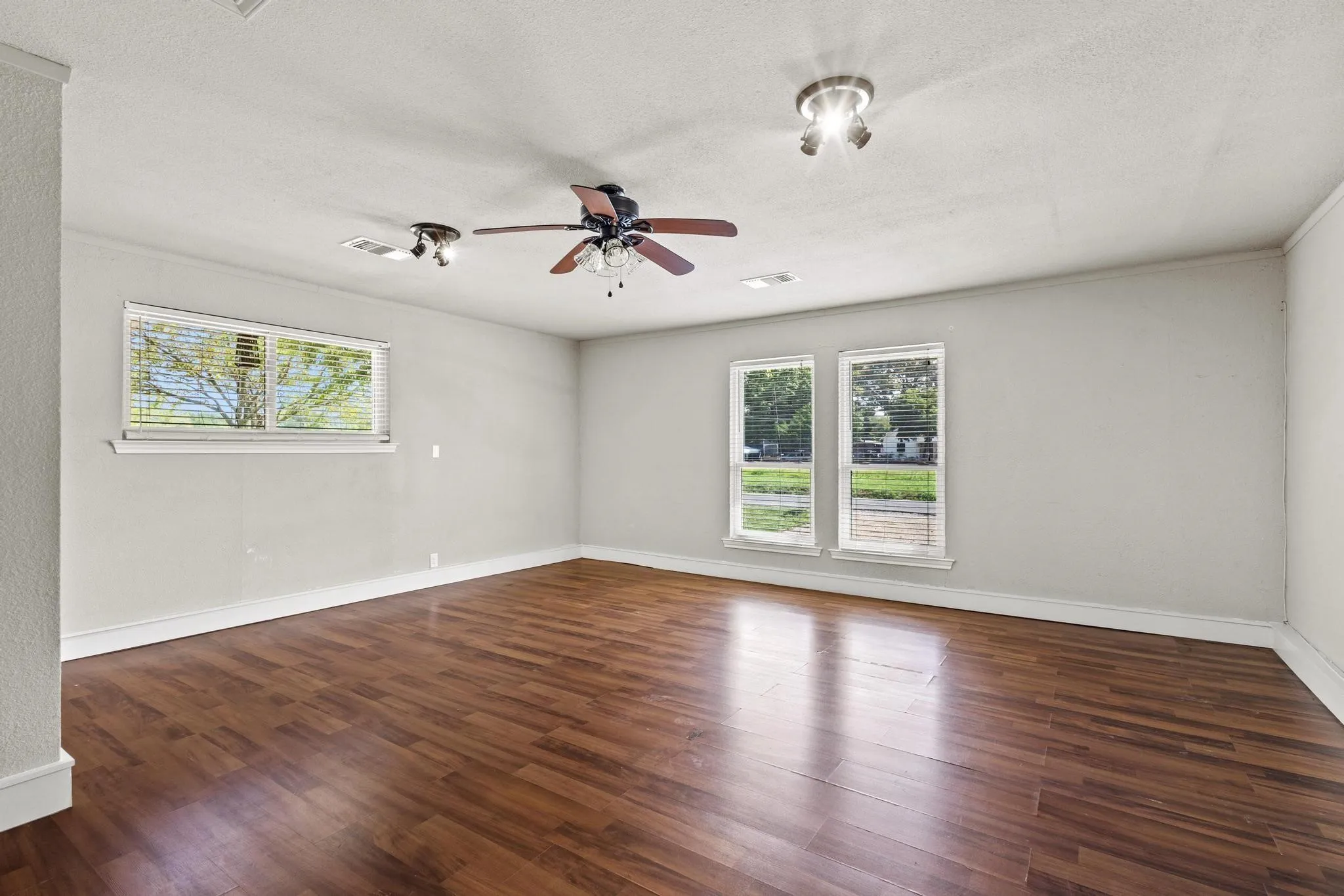 Spacious interior room featuring wood-finish flooring, light-toned walls, multiple windows, a ceiling fan, and track lighting fixtures