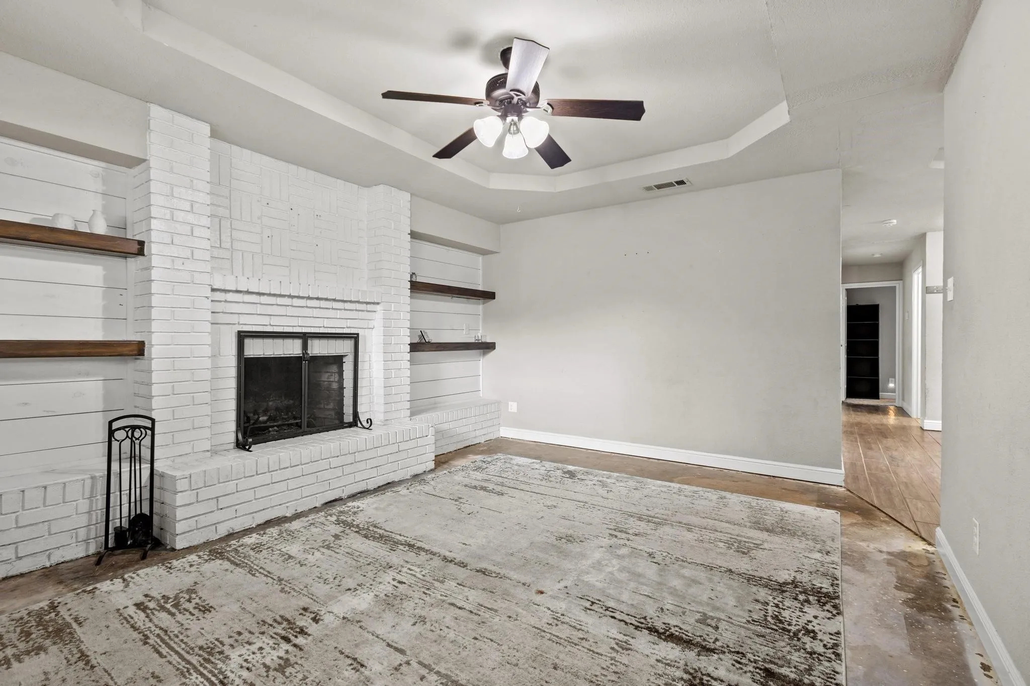 Living area featuring a white-painted brick fireplace with a black metal screen, integrated wood-finish floating shelves, a tray ceiling, and a ceiling fan. New carpet installed 08/12/2026.