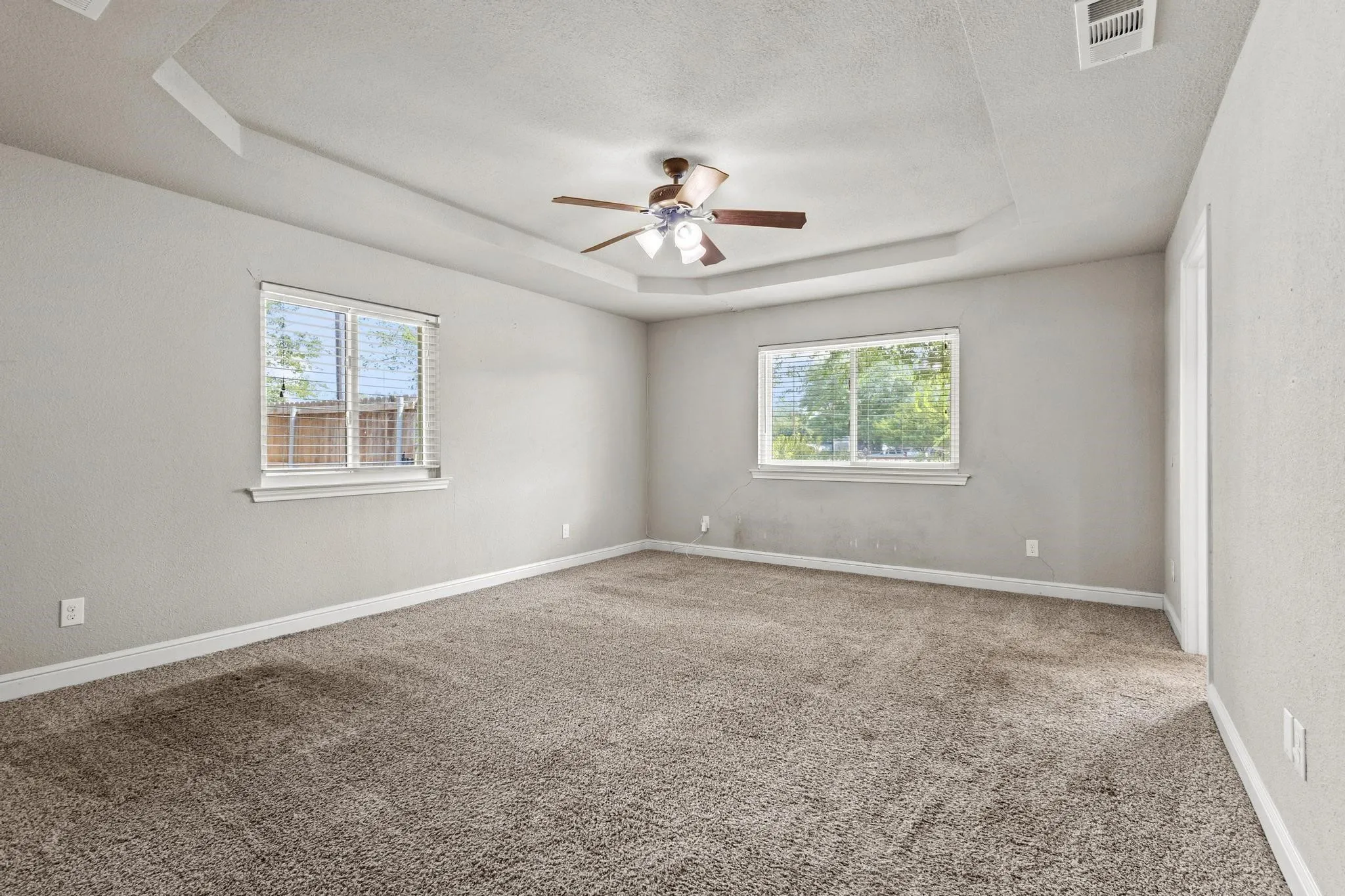 Spacious room featuring neutral carpeting, light gray wall tones, a tray ceiling with a mounted ceiling fan, and multiple windows with blinds. Carpet replaced 08-12-2026.