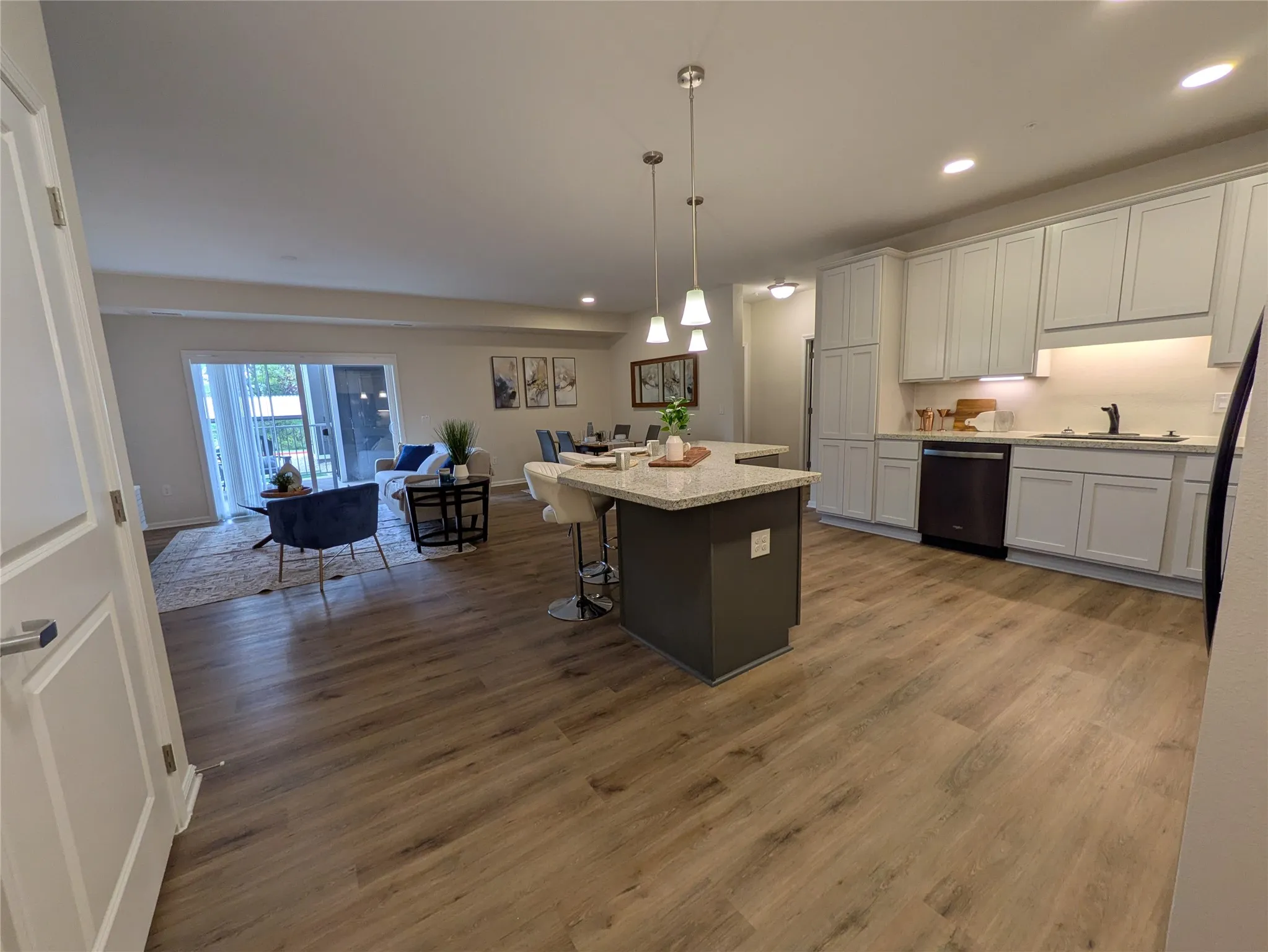 Open-concept living space featuring wood-finish flooring, a kitchen island with pendant lighting and bar seating, light-toned cabinetry, and a sliding glass door leading to an outdoor space