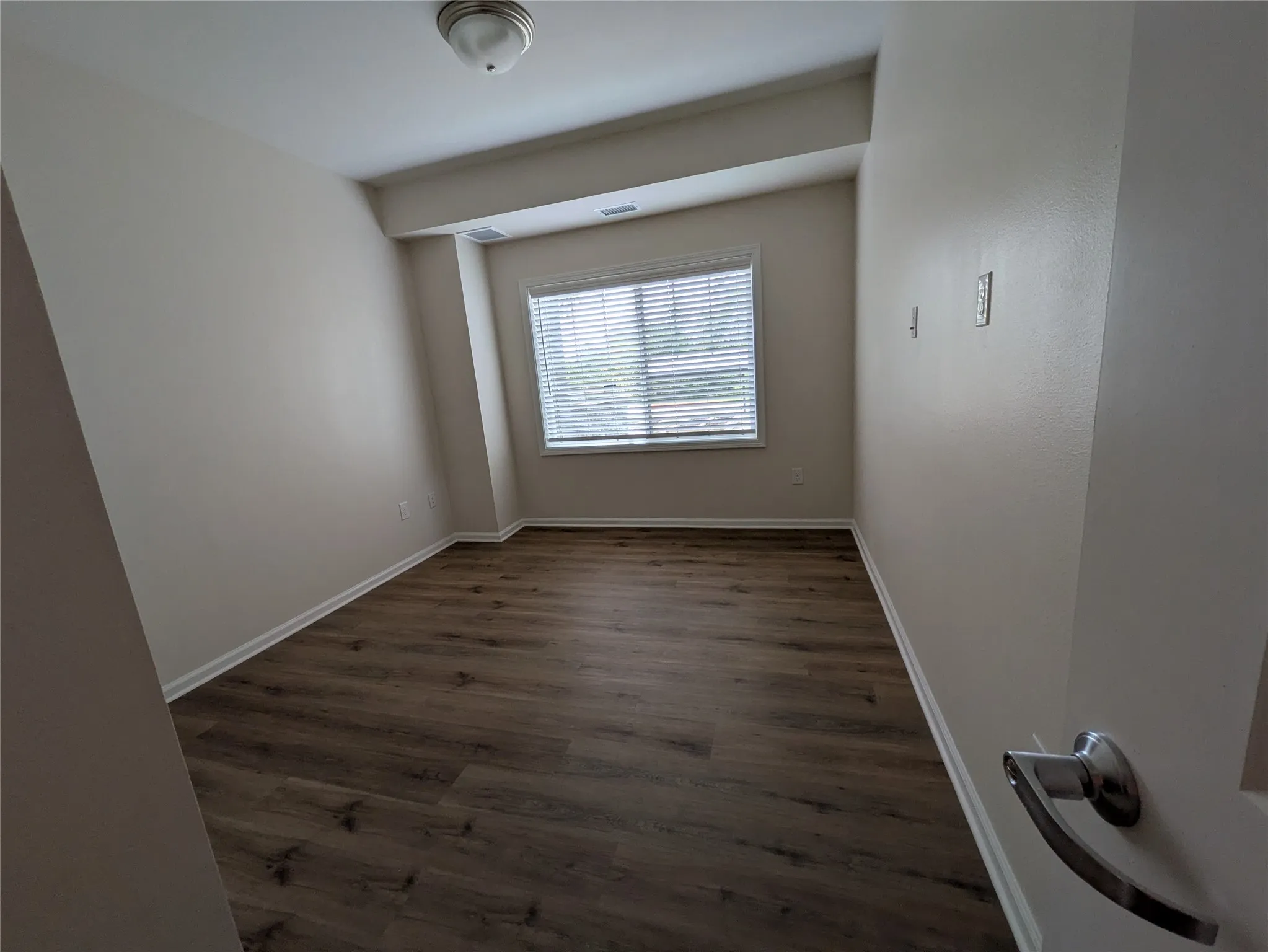Neutral-toned room featuring a single window with blinds, wood-finish flooring, and a flush-mount ceiling light
