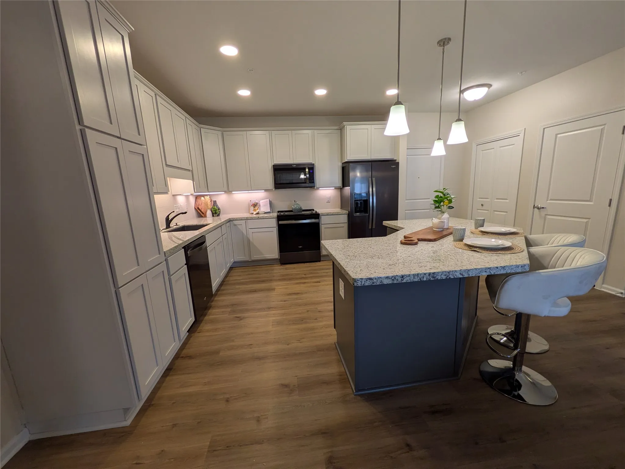 Contemporary kitchen featuring a multi-sided island with a light speckled countertop, wood-finish flooring, white shaker-style cabinetry, and recessed lighting
