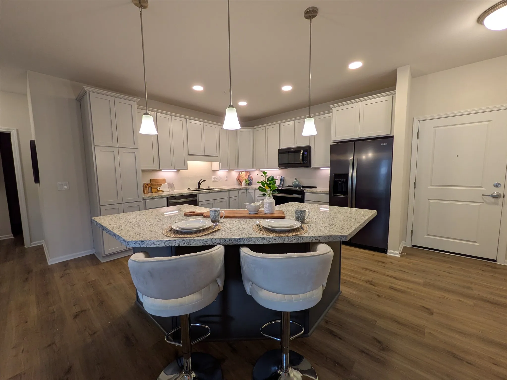 Open-concept kitchen featuring light-toned cabinetry, granite countertops, wood-finish flooring, a central island with pendant lighting, and stainless steel appliances