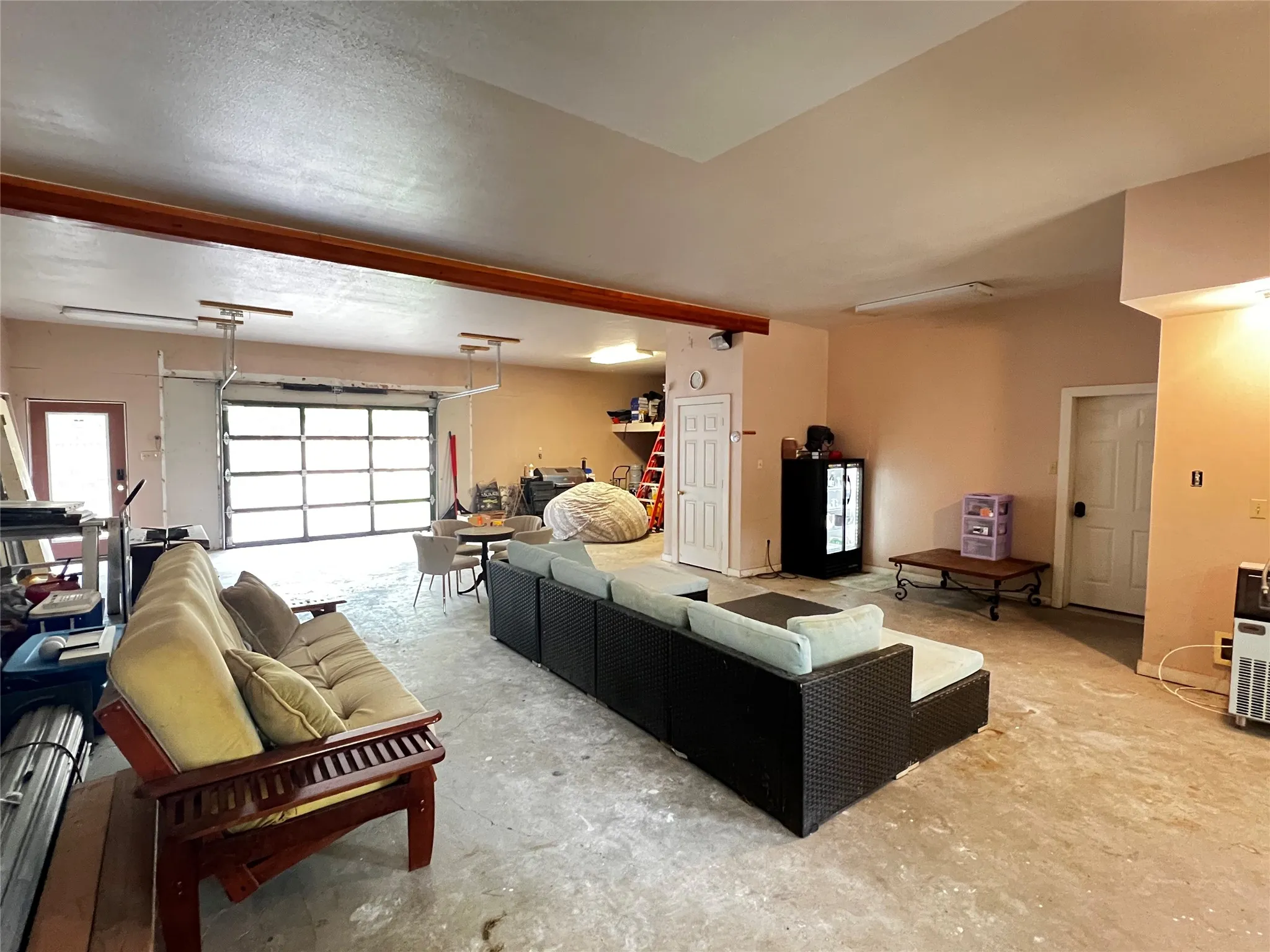 Spacious multi-purpose room featuring a large overhead glass-paneled garage door, exposed wood beam, concrete flooring, and multiple access doors
