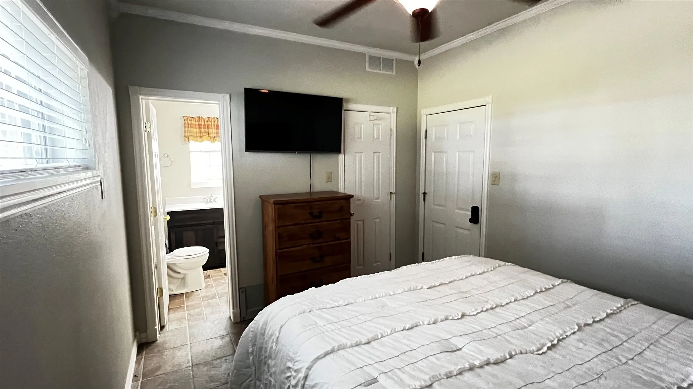 Primary bedroom with ensuite bathroom, ceiling fan, wall-mounted television, and wood-finish flooring