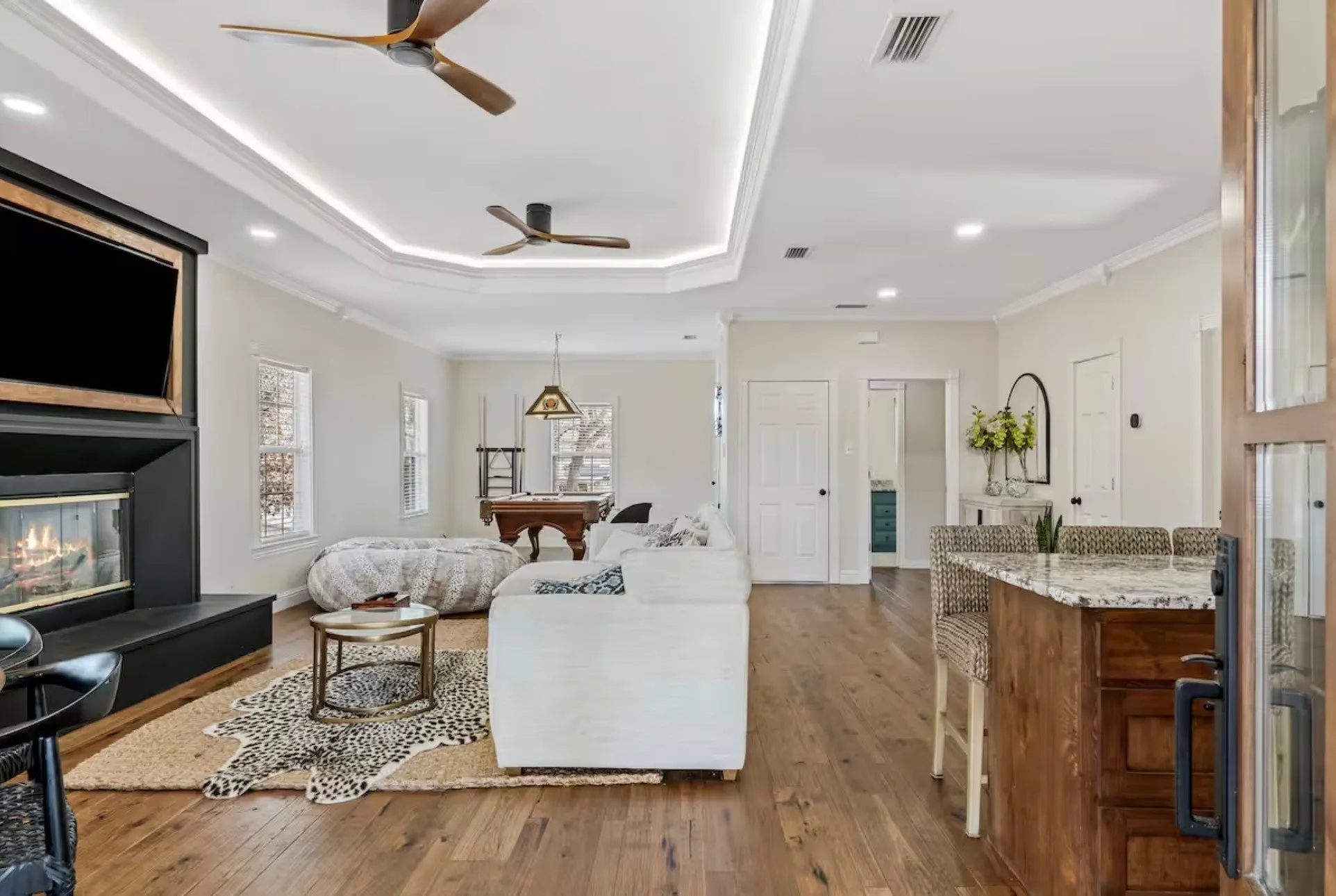 Spacious living area featuring wood-finish flooring, a tray ceiling with recessed lighting and two ceiling fans, a built-in fireplace with a dark mantel, and a kitchen island with a light-toned countertop