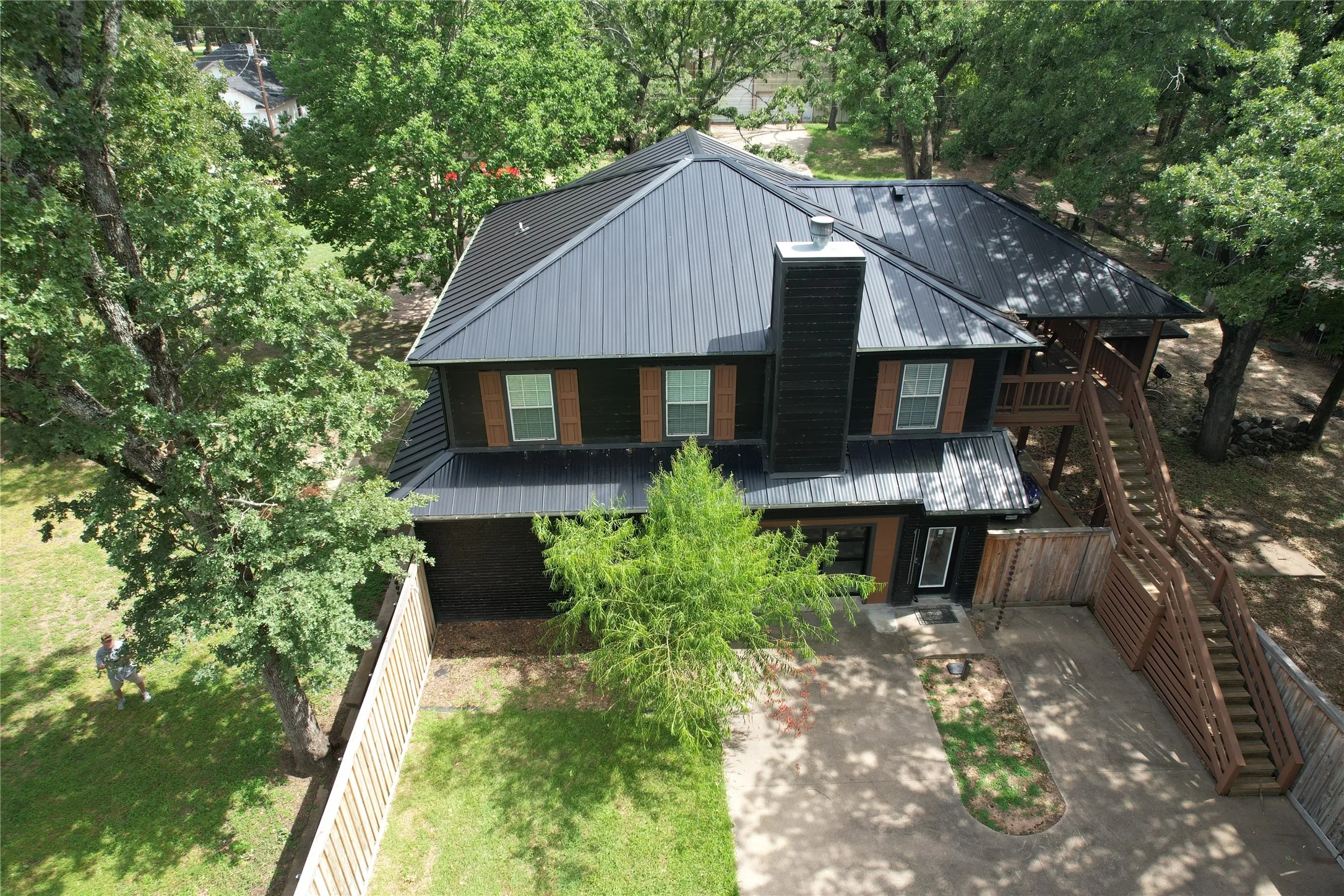 Aerial view of the White Oak house elevation featuring the metal roof, full glass roll-up garage door, entry door, grass yard, ample driveway space, and easy walk-up stairs to upper deck with lake view and main second level living area.