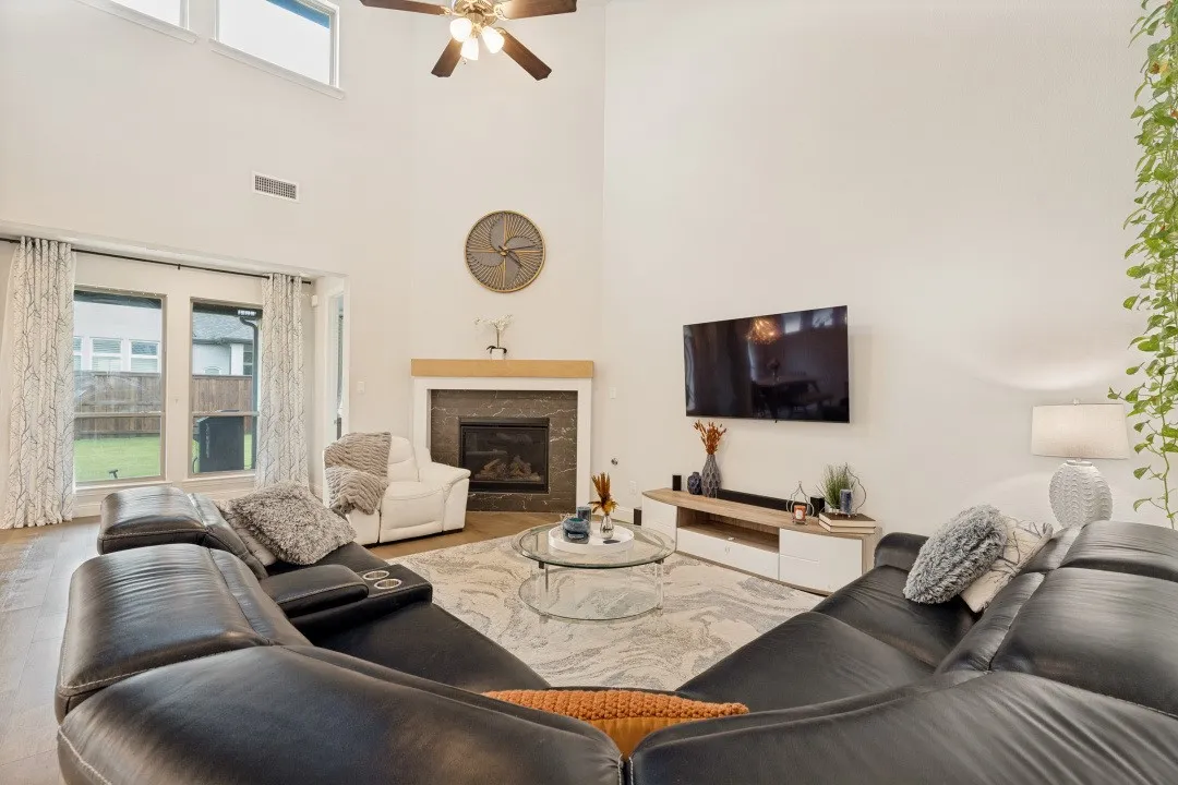 Vaulted ceiling living room featuring a ceiling fan, fireplace with a wood mantel, and large windows
