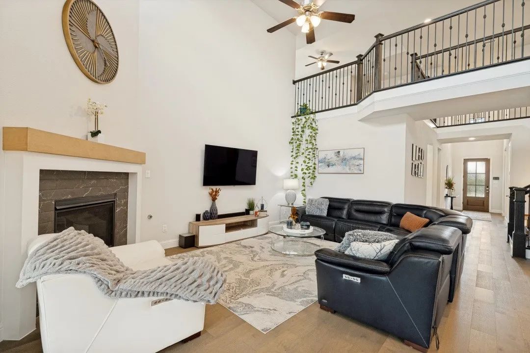 Spacious living area featuring high vaulted ceilings, a built-in fireplace with a stone surround, wood-finish flooring, an open upper-level walkway with wrought iron railings, and a prominent front entry door with a glass insert