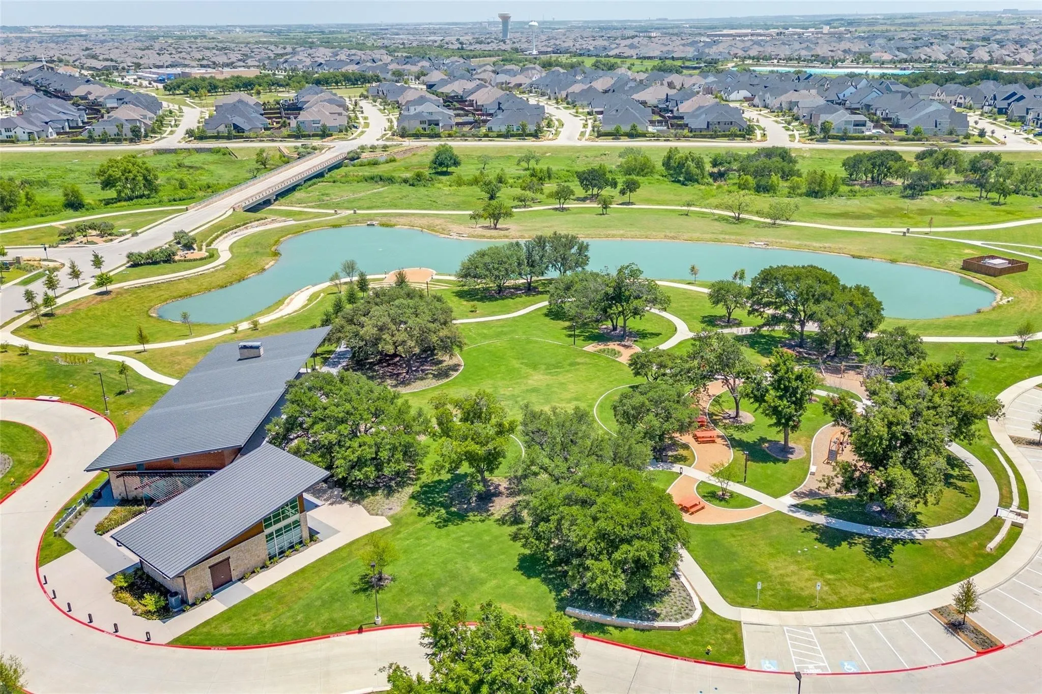 Expansive community greenspace featuring a large pond, walking paths, and a modern recreation building with a dark metal roof