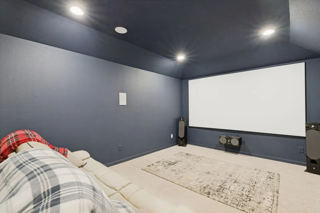 Dedicated media room featuring a large projection screen, recessed lighting, wall-mounted center speaker, carpeted flooring, and dark-toned walls