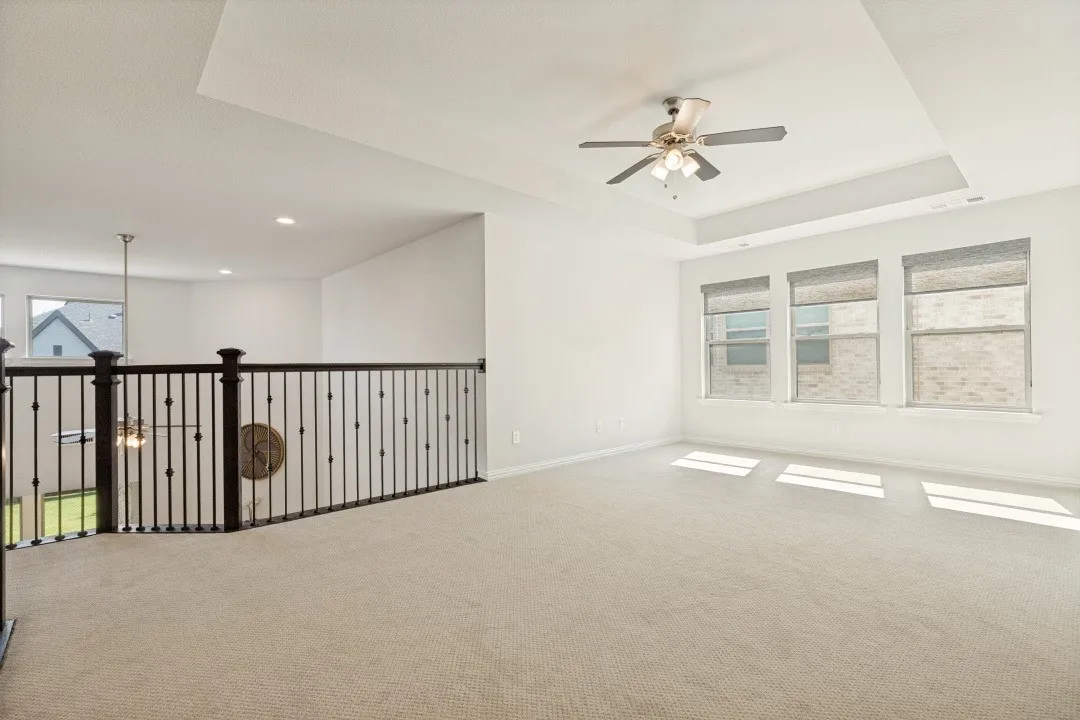 Carpeted loft area featuring a tray ceiling with a flush-mount fan, three windows with roller shades, and a dark iron spindle railing