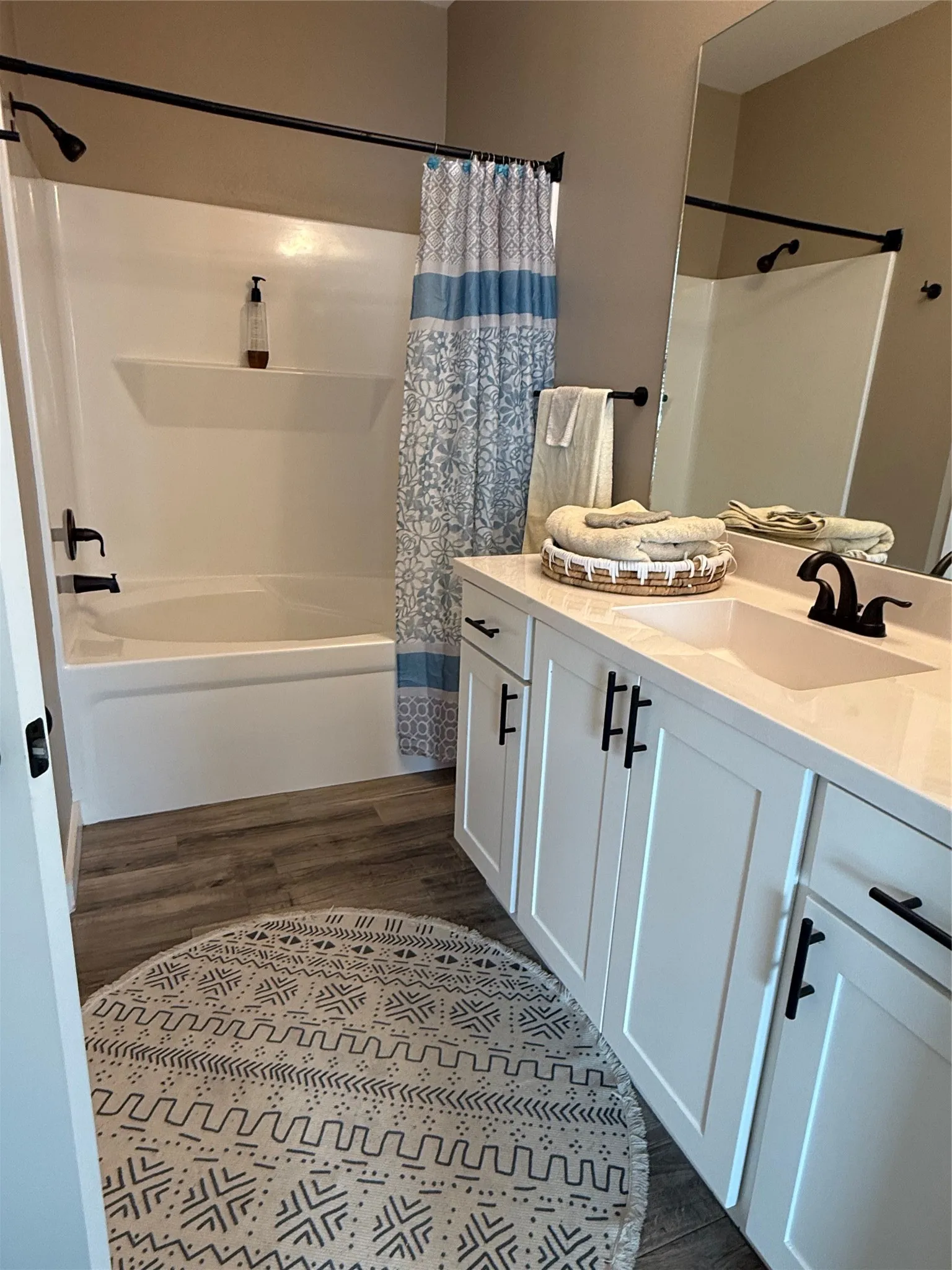 Bathroom featuring a white bathtub/shower combination with matte black fixtures, a dual-sink vanity with white cabinetry and matte black hardware, a large vanity mirror, and wood-finish flooring