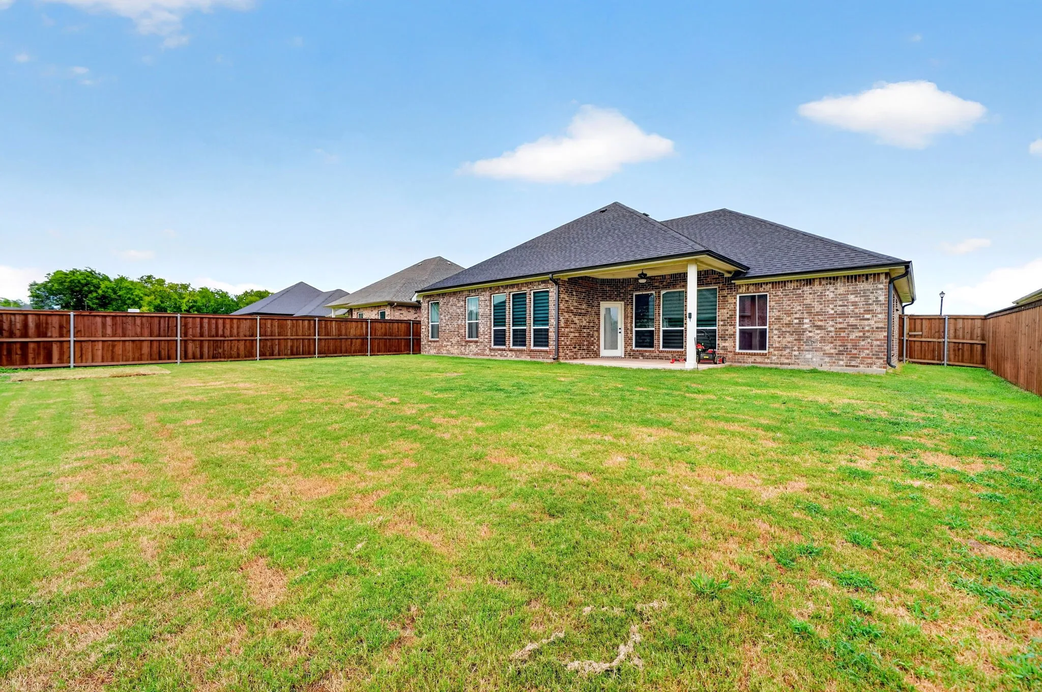 Expansive backyard featuring a privacy fence, covered patio, brick exterior, multiple windows, and a dark shingle roof