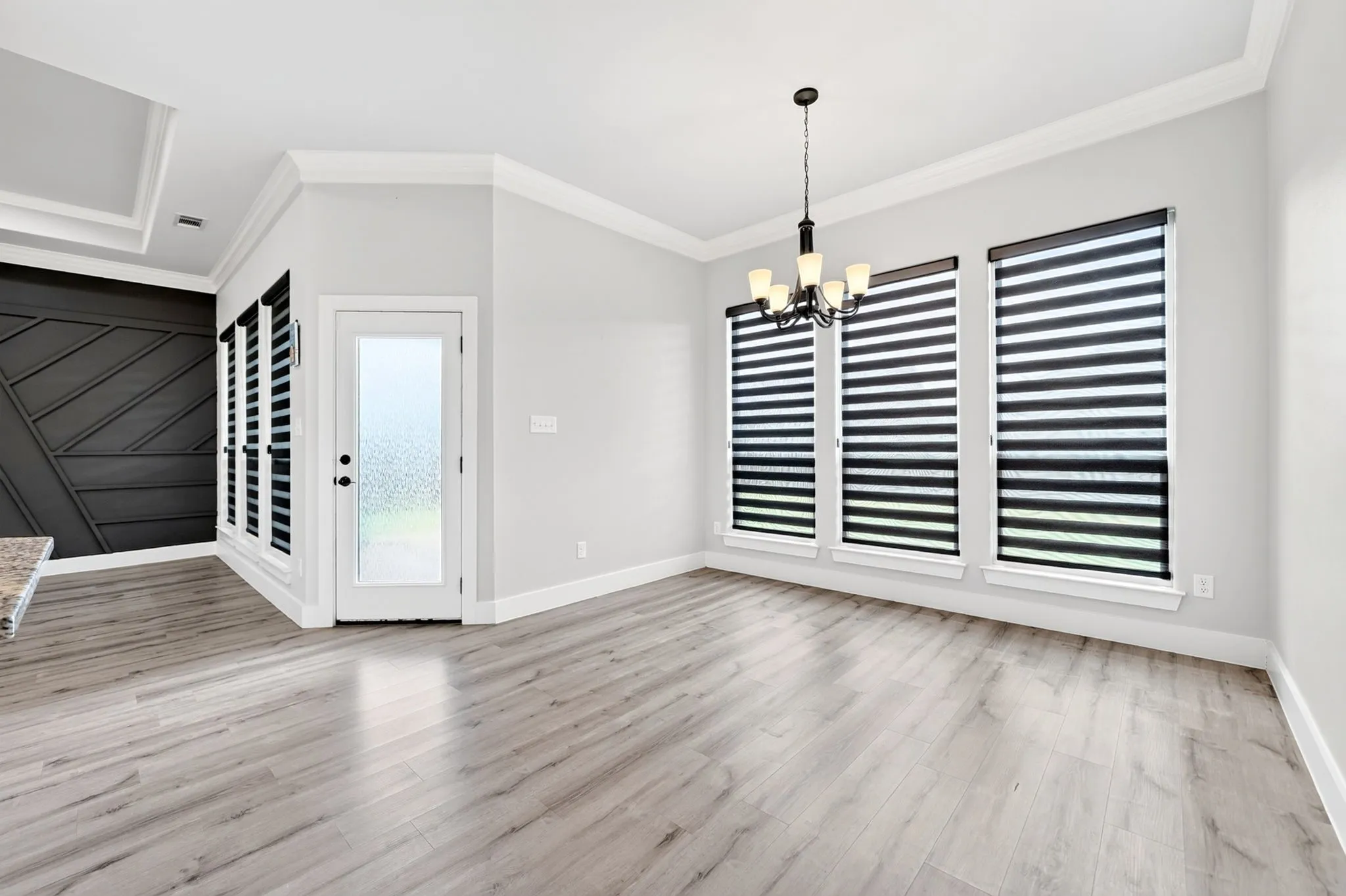 Spacious room with light wood-finish flooring, featuring a dark accent wall with geometric paneling, multiple windows with horizontal blinds, and a frosted glass panel door