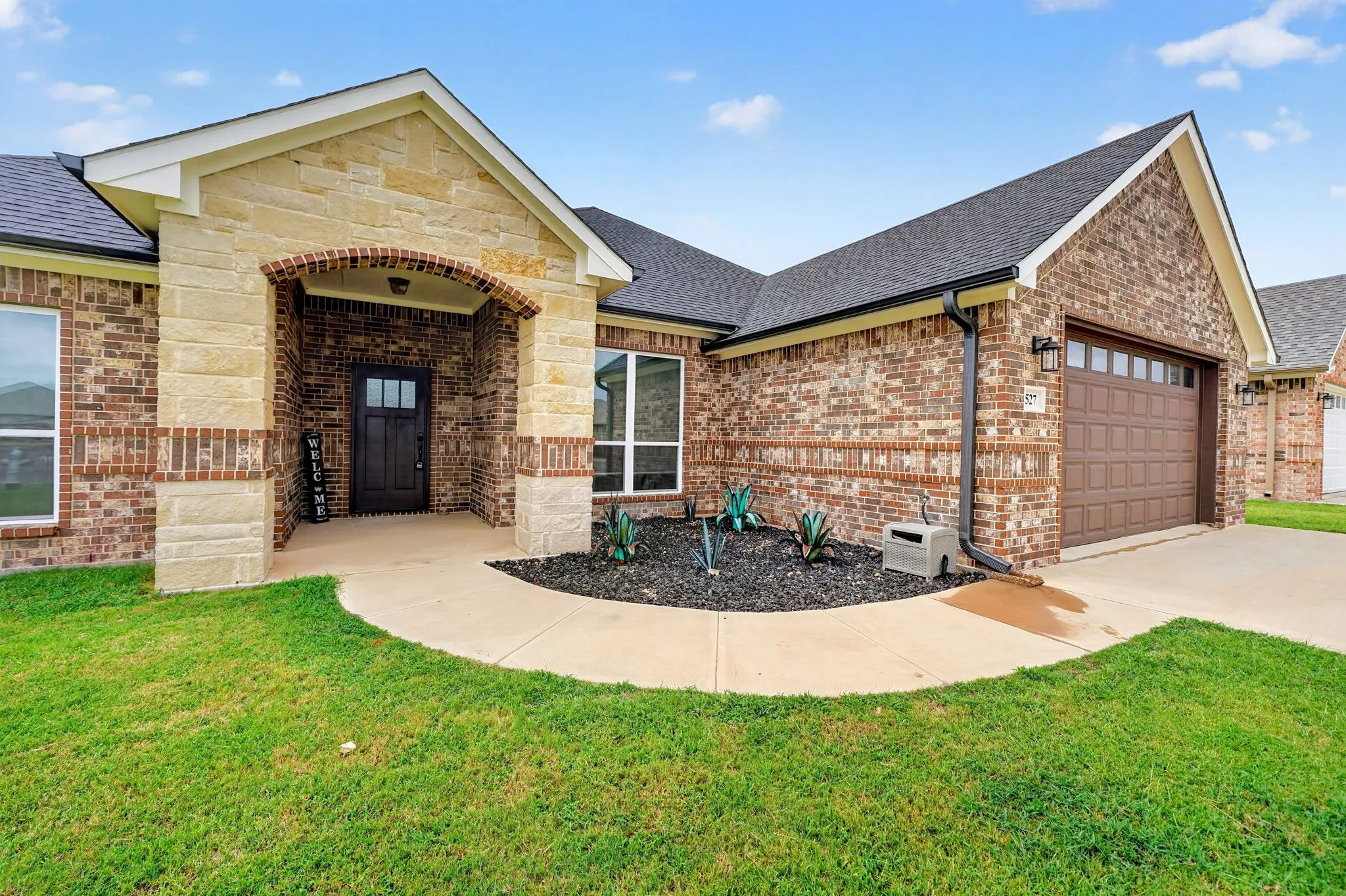 Brick and stone exterior featuring a covered entry porch, a dark wood-finish front door with glass inserts, a landscaped planting bed, and an attached garage with a panel door