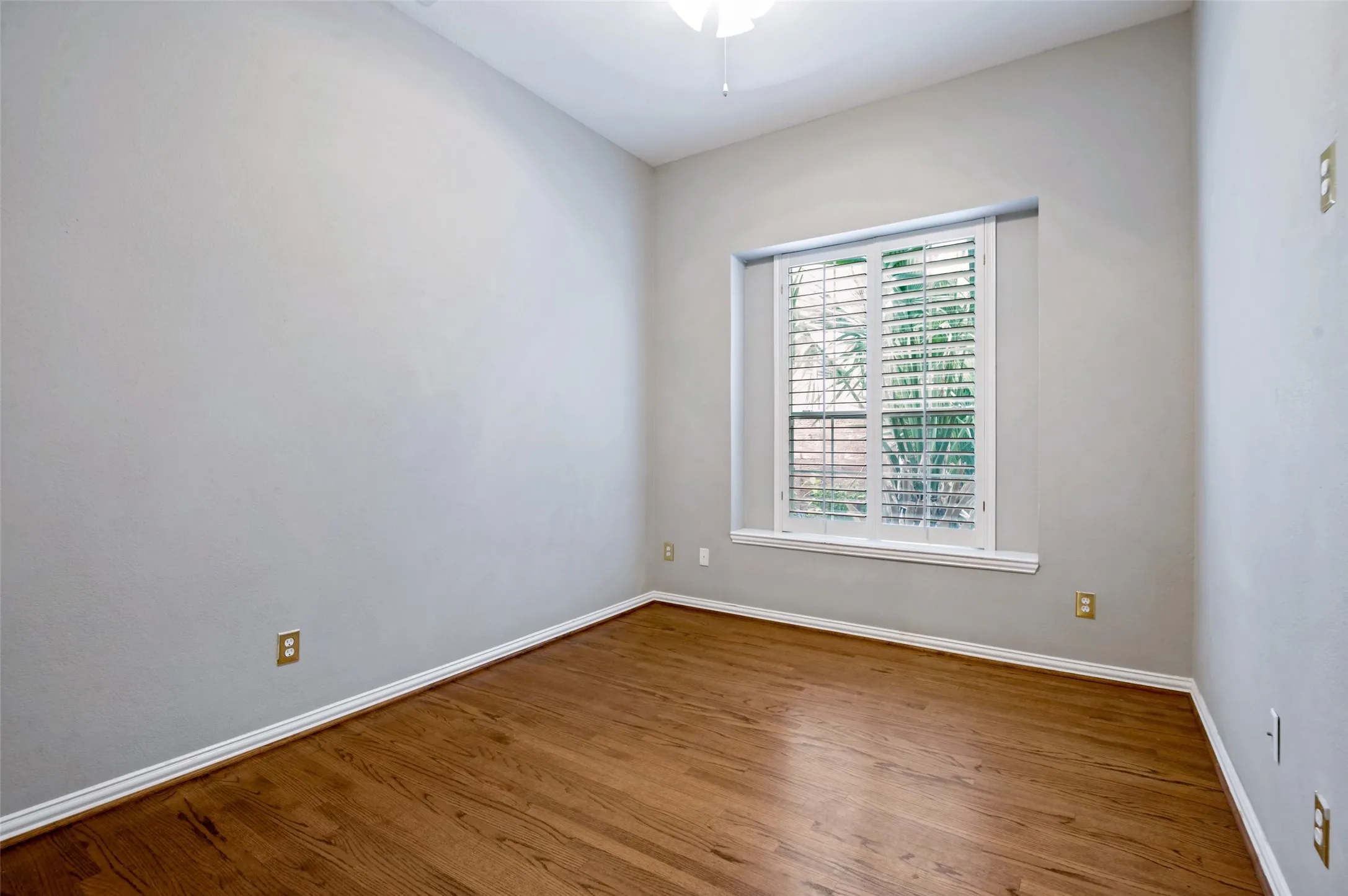 Room featuring wood-finish flooring, white baseboards, a window with plantation shutters, light gray wall paint, and a ceiling fan