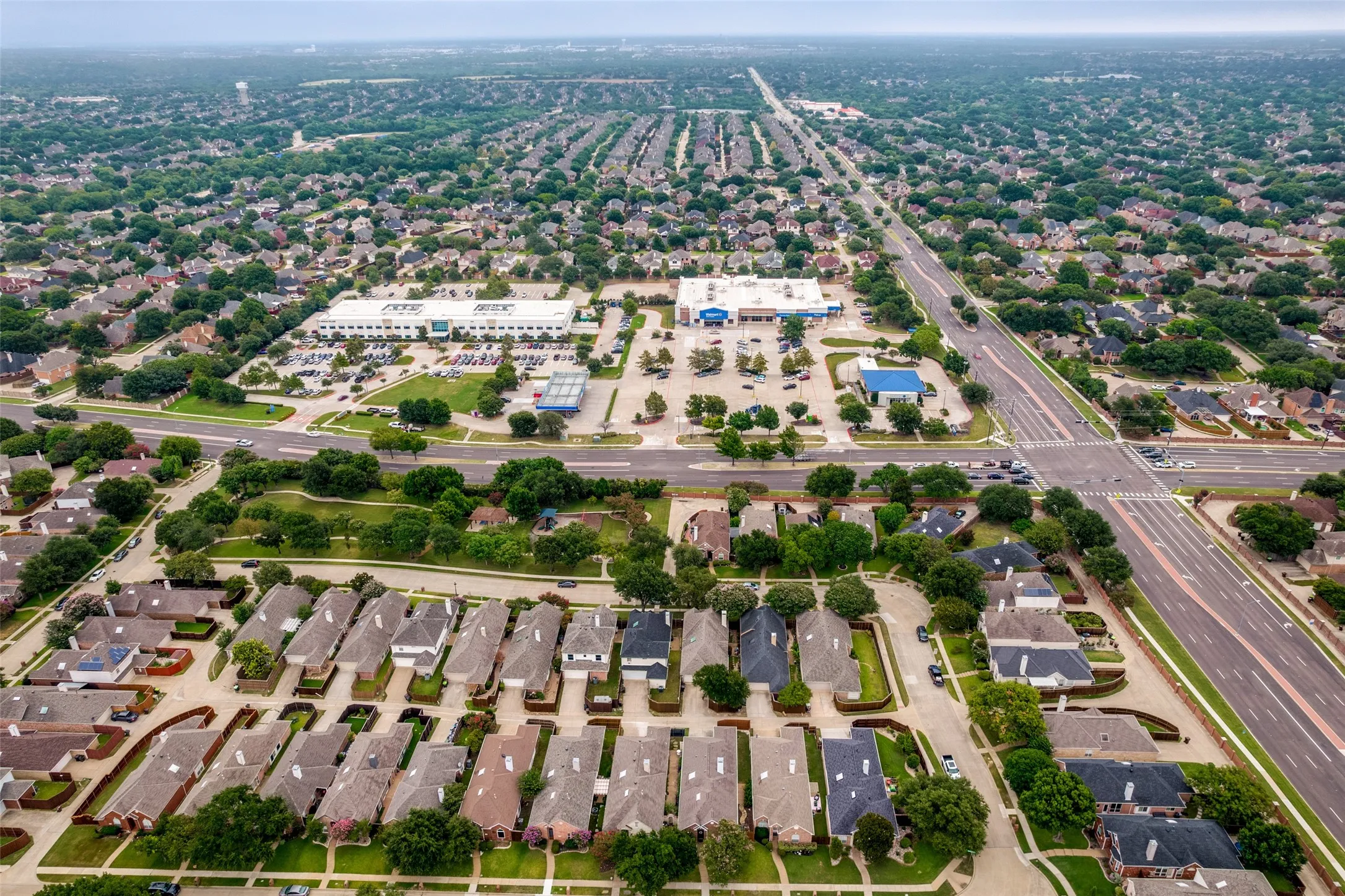 Expansive aerial perspective showcasing a residential neighborhood with numerous single-family homes, mature trees, and a commercial area featuring retail buildings and parking