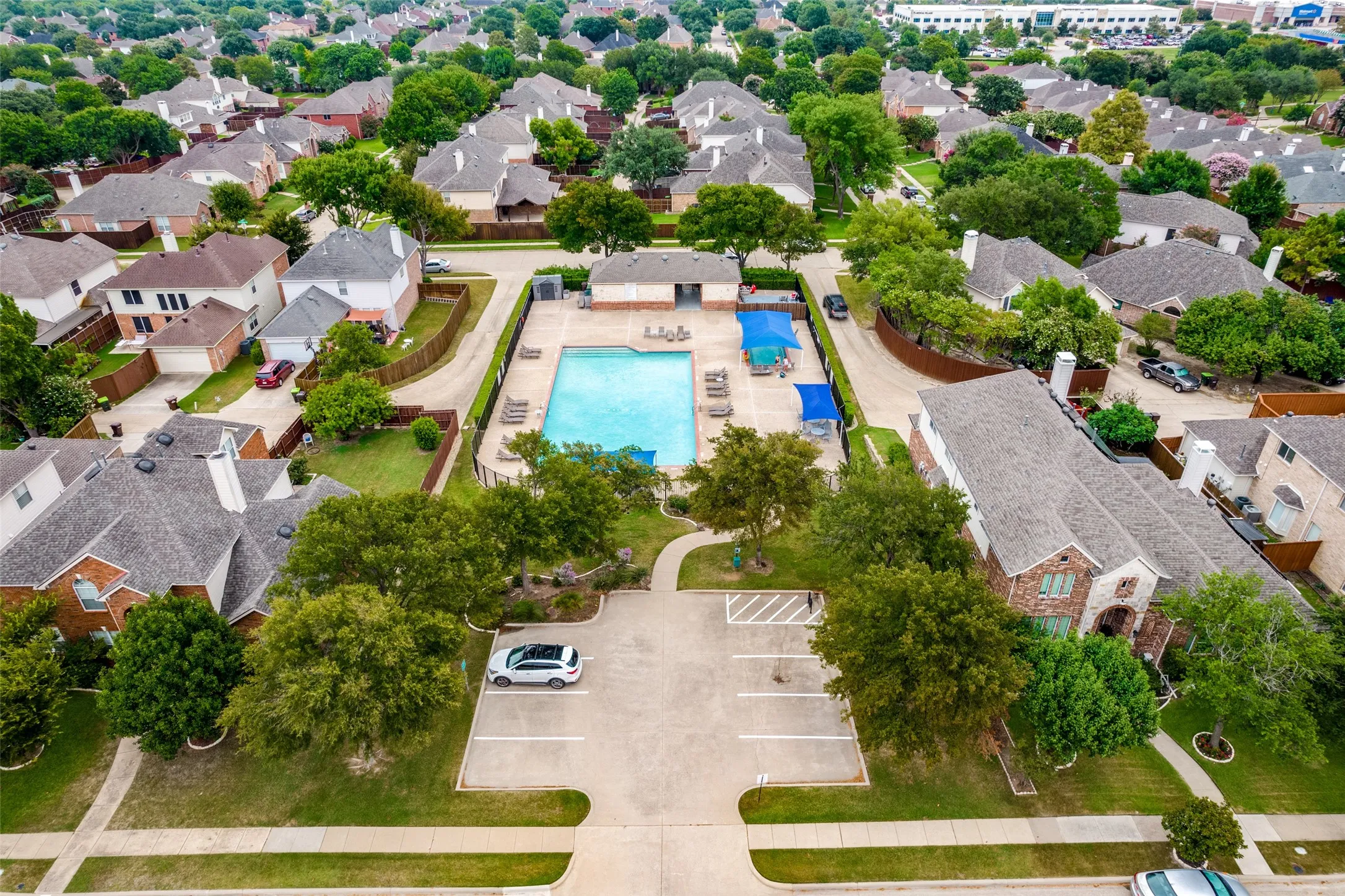 Community swimming pool with surrounding lounge areas and sun shades, bordered by mature trees and a dedicated parking lot
