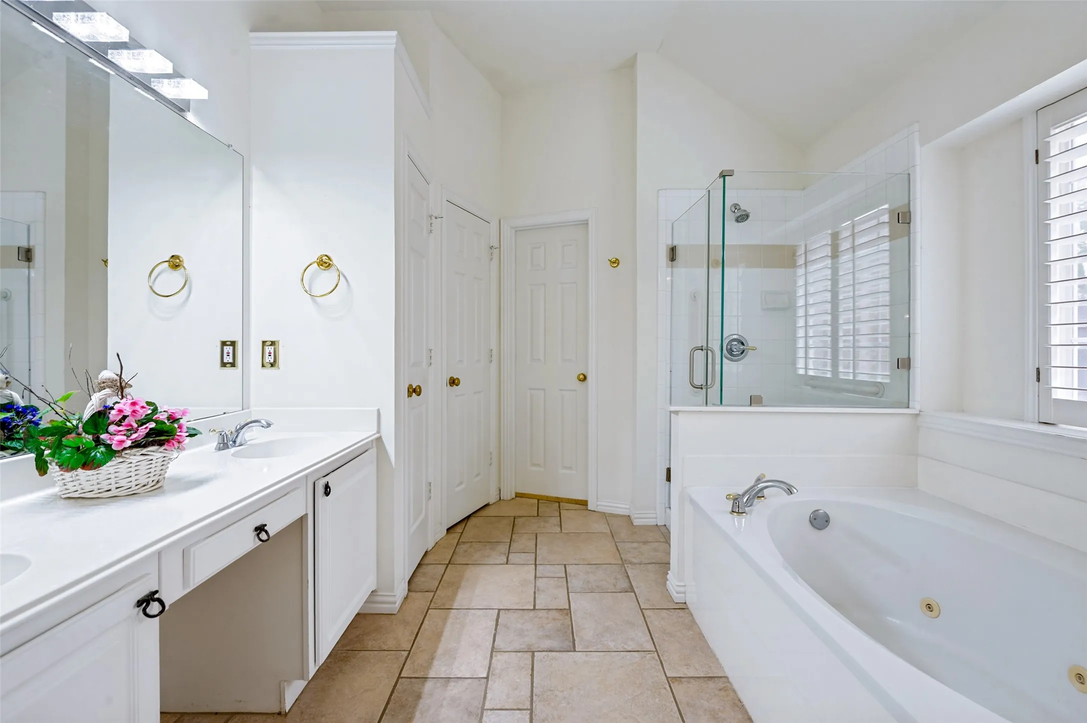 Spacious bathroom featuring a dual vanity, a built-in jetted tub, a glass-enclosed shower, wood shutters, and tile flooring