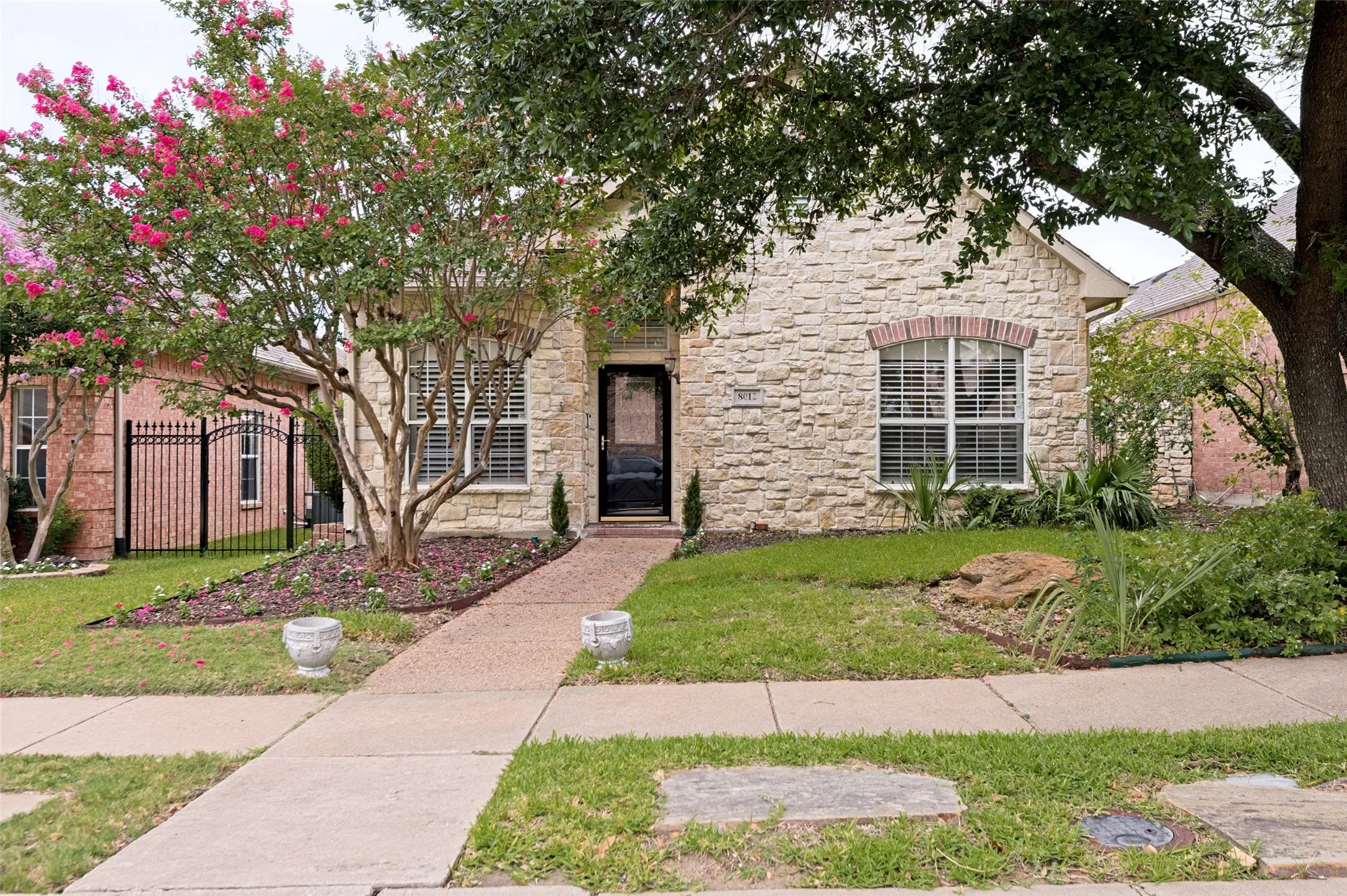 Stone facade residence featuring a dark paneled entry door, arched windows with shutters, and mature landscaping with flowering trees
