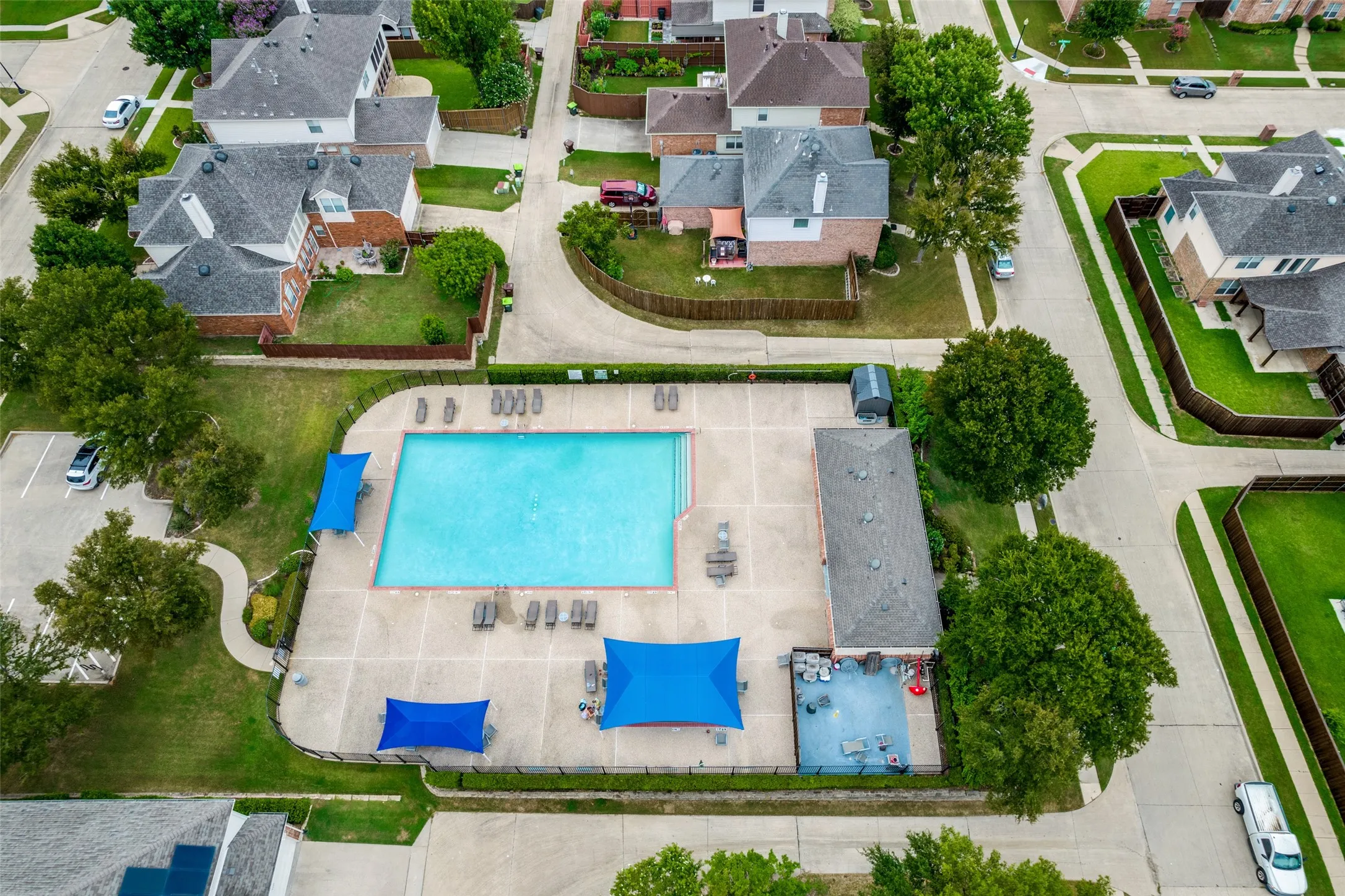 Community swimming pool featuring a large rectangular pool, expansive concrete deck, multiple lounge chairs, blue shade structures, and a dedicated pool house