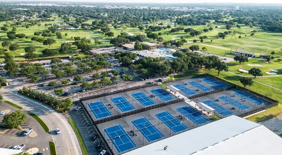 Premier sports facility featuring multiple professional-grade hard courts with blue playing surfaces, expansive covered pavilion structure, and manicured grounds surrounded by mature​​‌​​​​‌​​‌‌​‌‌​​​‌‌​‌​‌​‌​​​‌​​ landscaping