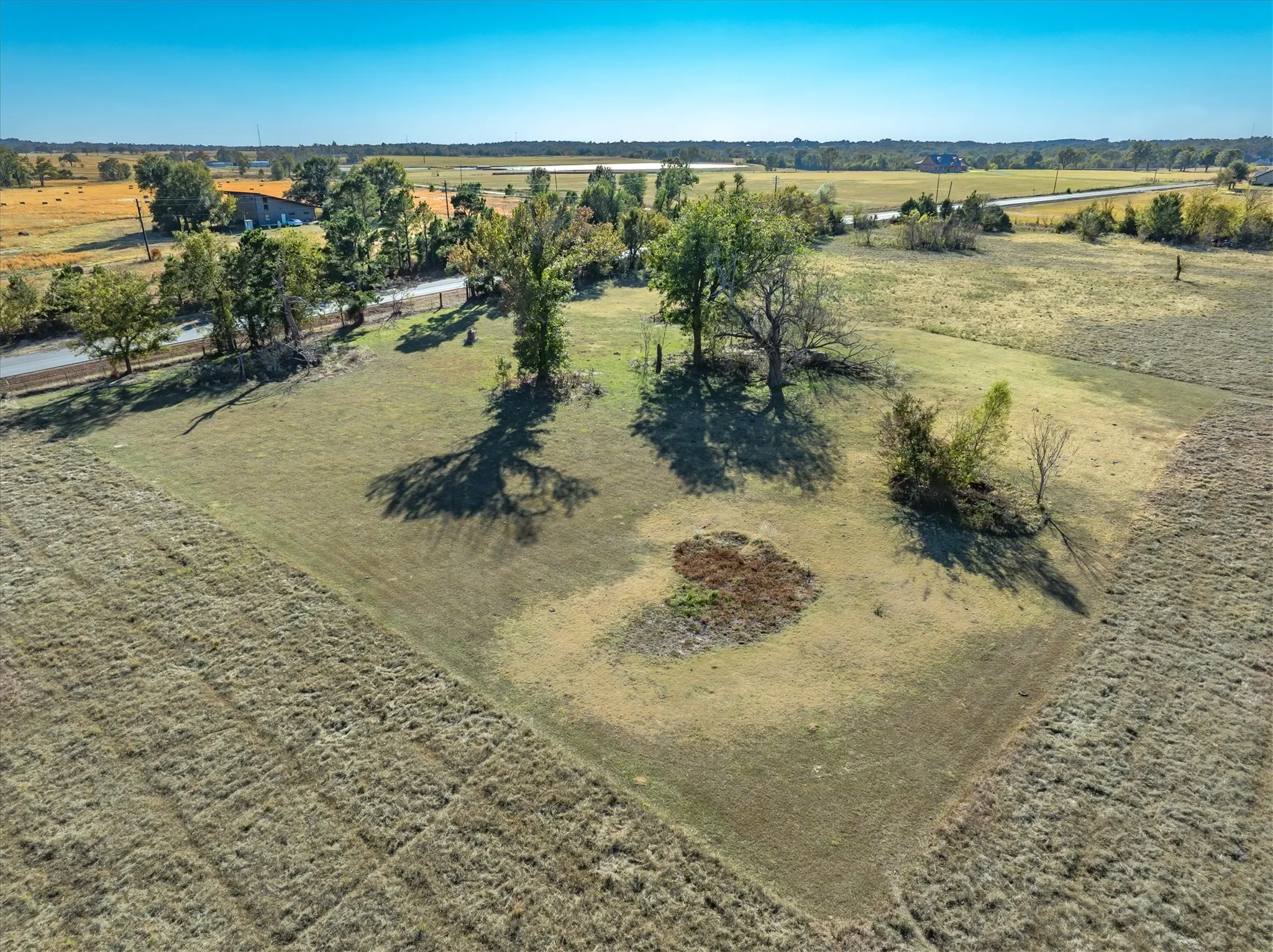 Expansive parcel featuring a defined grassy area, surrounding fields, mature trees, and clear blue skies