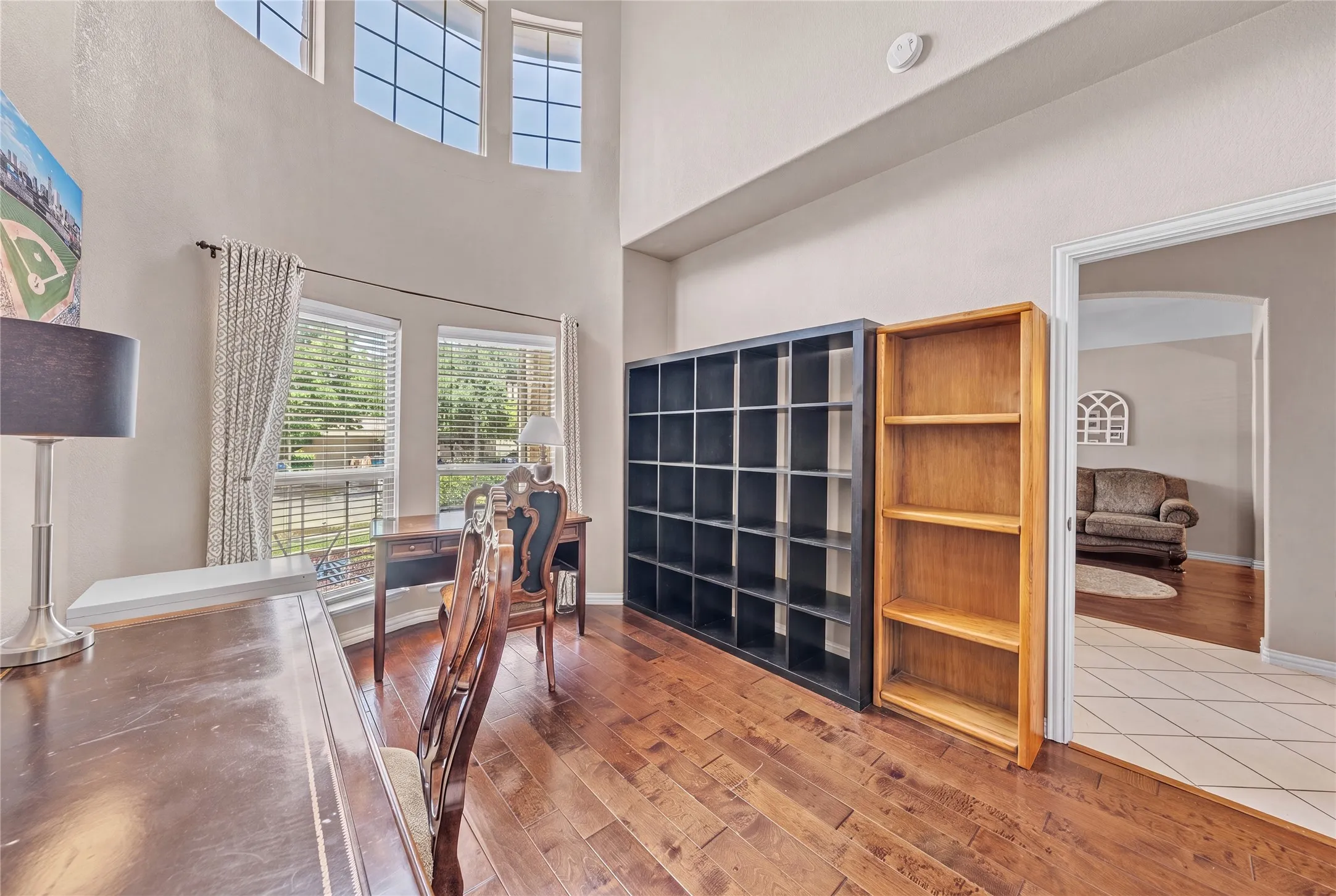 Room with wood-finish flooring, featuring a high ceiling, upper transom windows, and lower windows with blinds