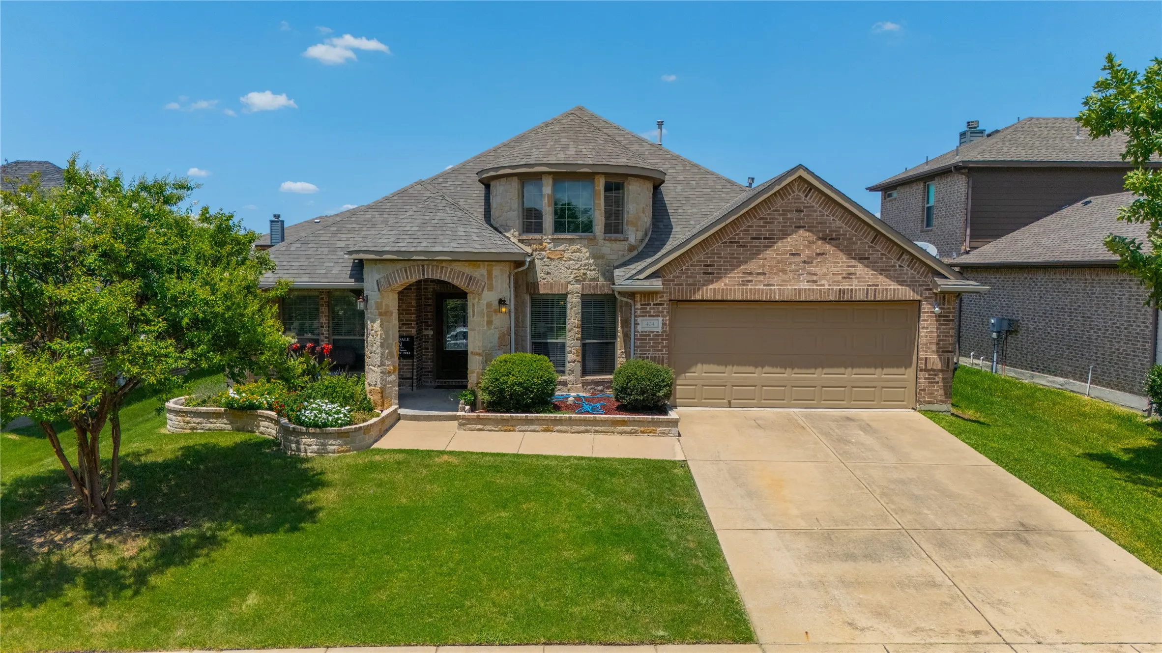 Brick and stone facade residence featuring a two-car garage, arched entry porch, and a stone-edged garden bed with mature landscaping