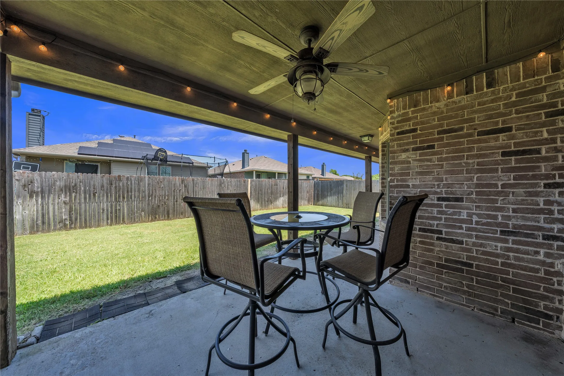 Covered outdoor patio featuring a ceiling fan with integrated lighting, brick accent wall, string lighting, and concrete flooring
