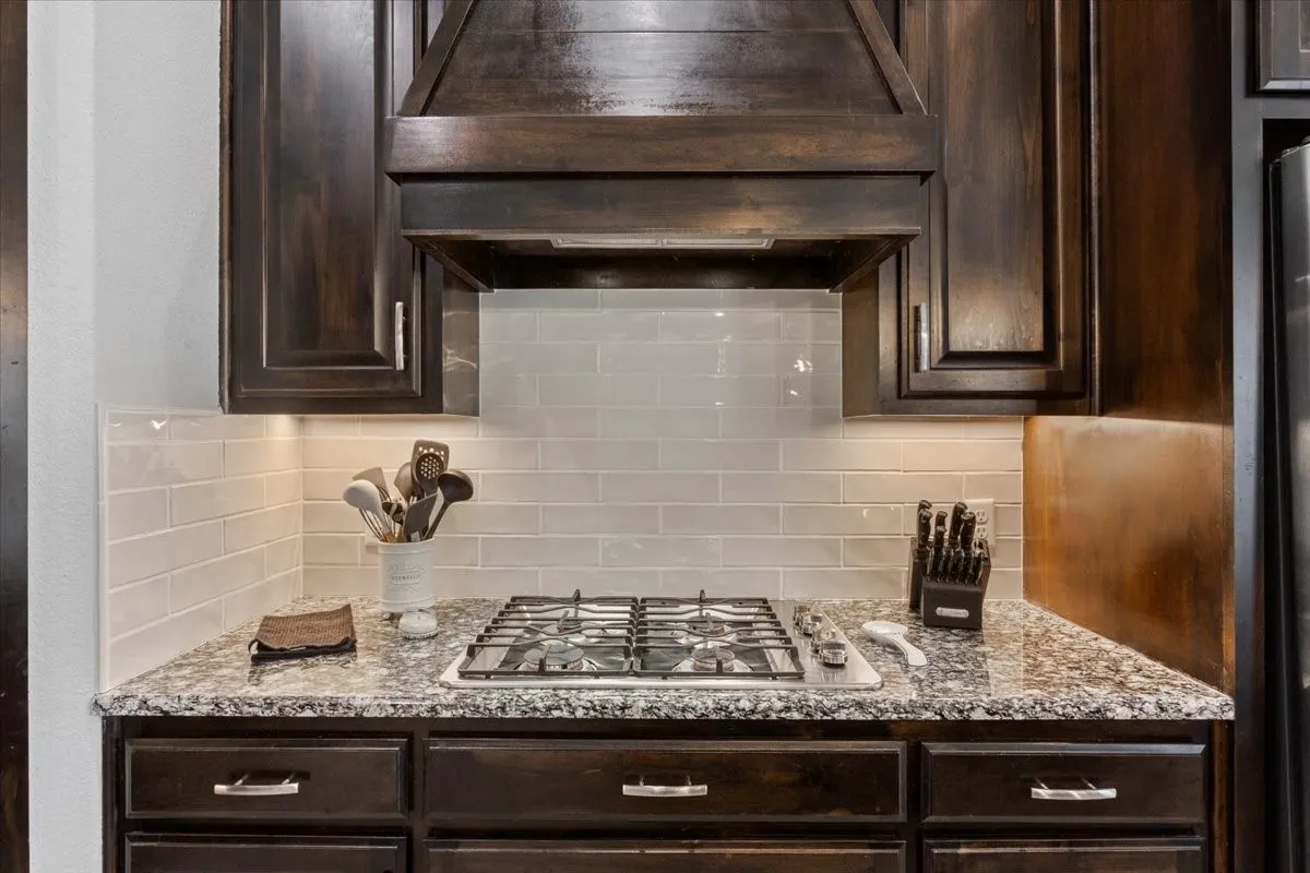 Kitchen workspace featuring a built-in gas range, granite-finish countertop, subway tile backsplash, dark wood cabinetry, and under-cabinet lighting