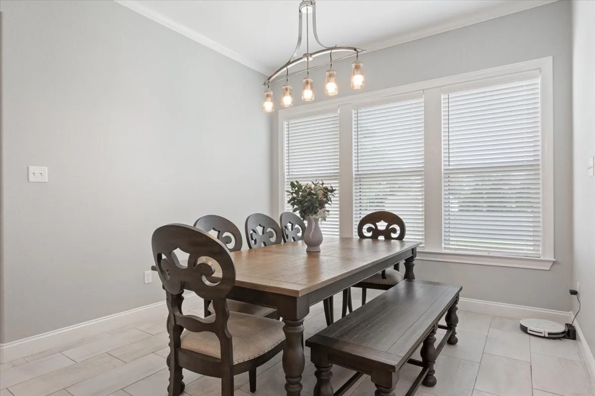 Bright dining area featuring a linear chandelier with Edison bulbs, multiple windows with blinds, light tile flooring, and crisp white baseboards and crown molding