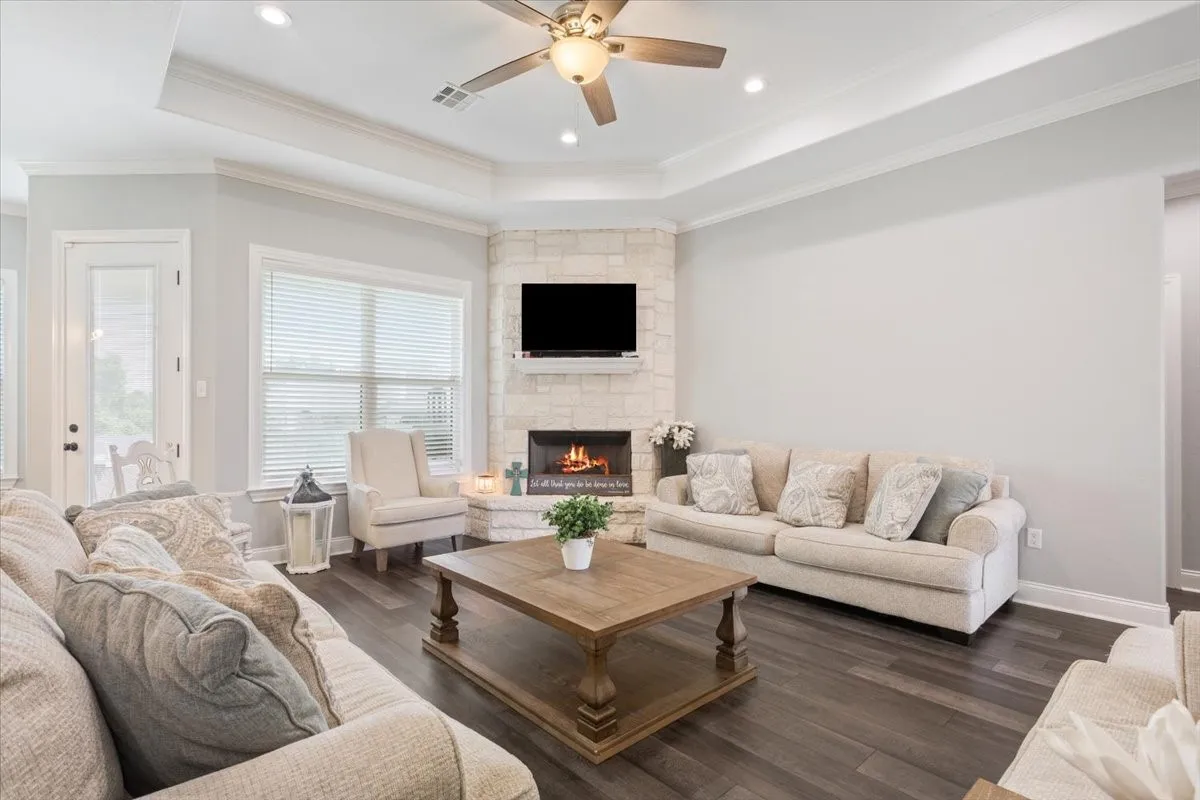 Living area featuring a corner stone fireplace, wood-finish flooring, recessed lighting, a ceiling fan, and a tray ceiling