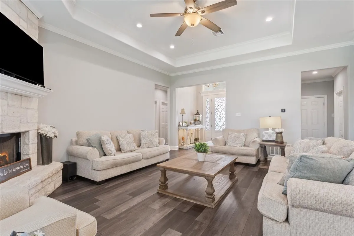 Living area featuring a stone-clad fireplace with mantel, dark wood-finish flooring, a tray ceiling with recessed lighting, and a ceiling fan