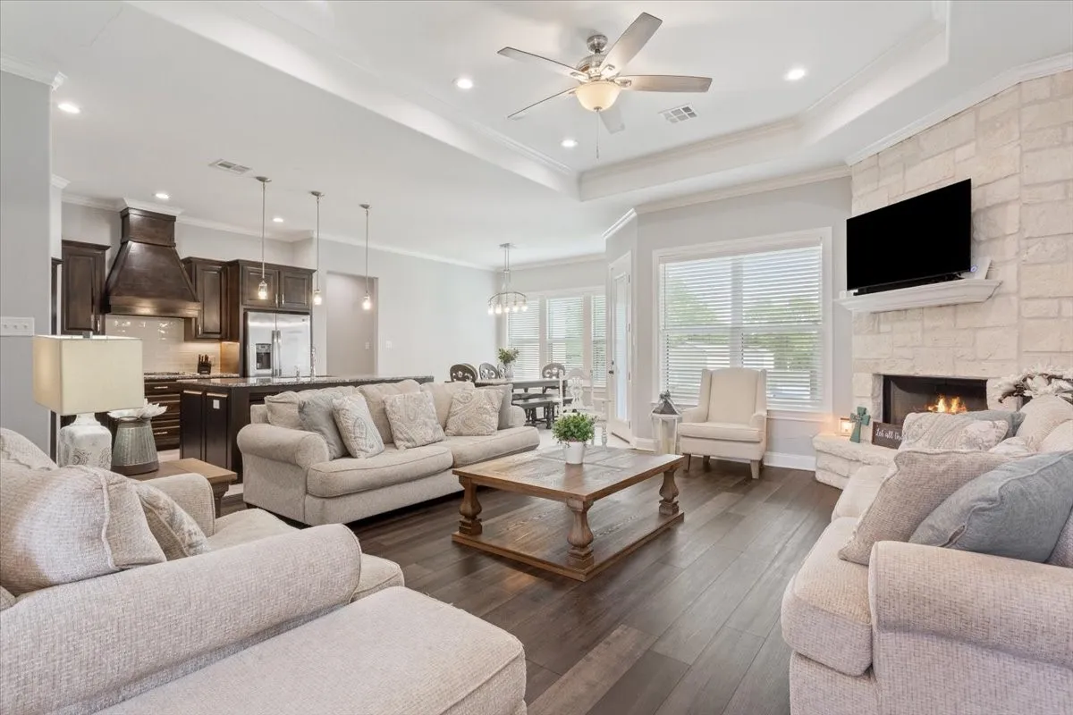 Open-concept living area featuring rich wood-finish flooring, a stone-clad fireplace, and an integrated kitchen with dark cabinetry and pendant lighting