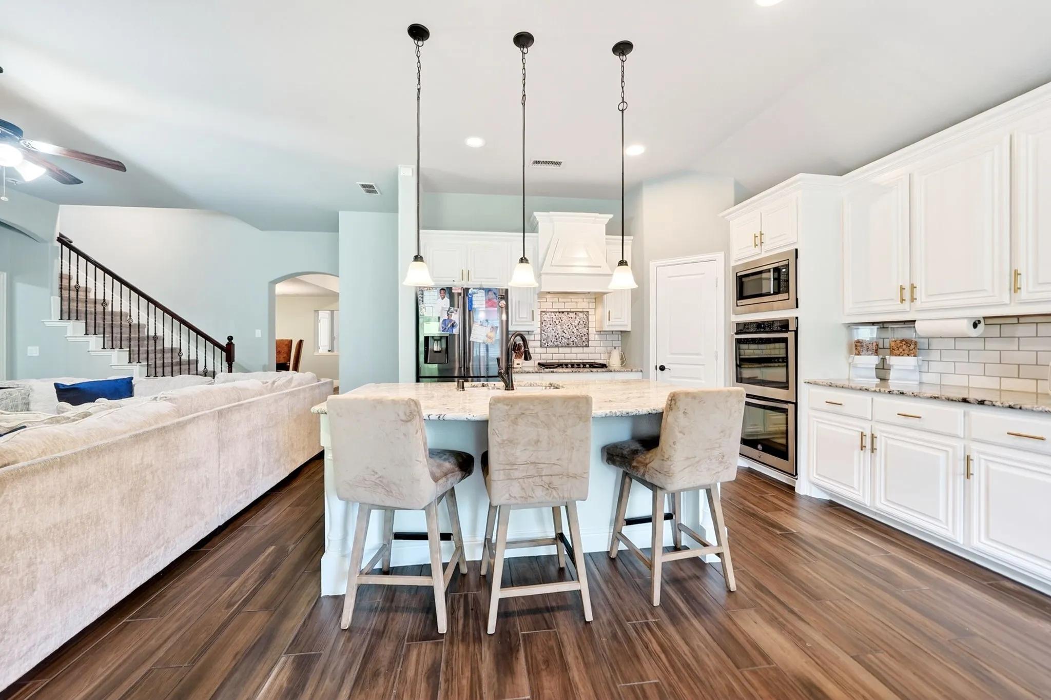 Spacious kitchen featuring an island with pendant lighting, white cabinetry with gold-tone hardware, built-in double ovens, and wood-finish flooring