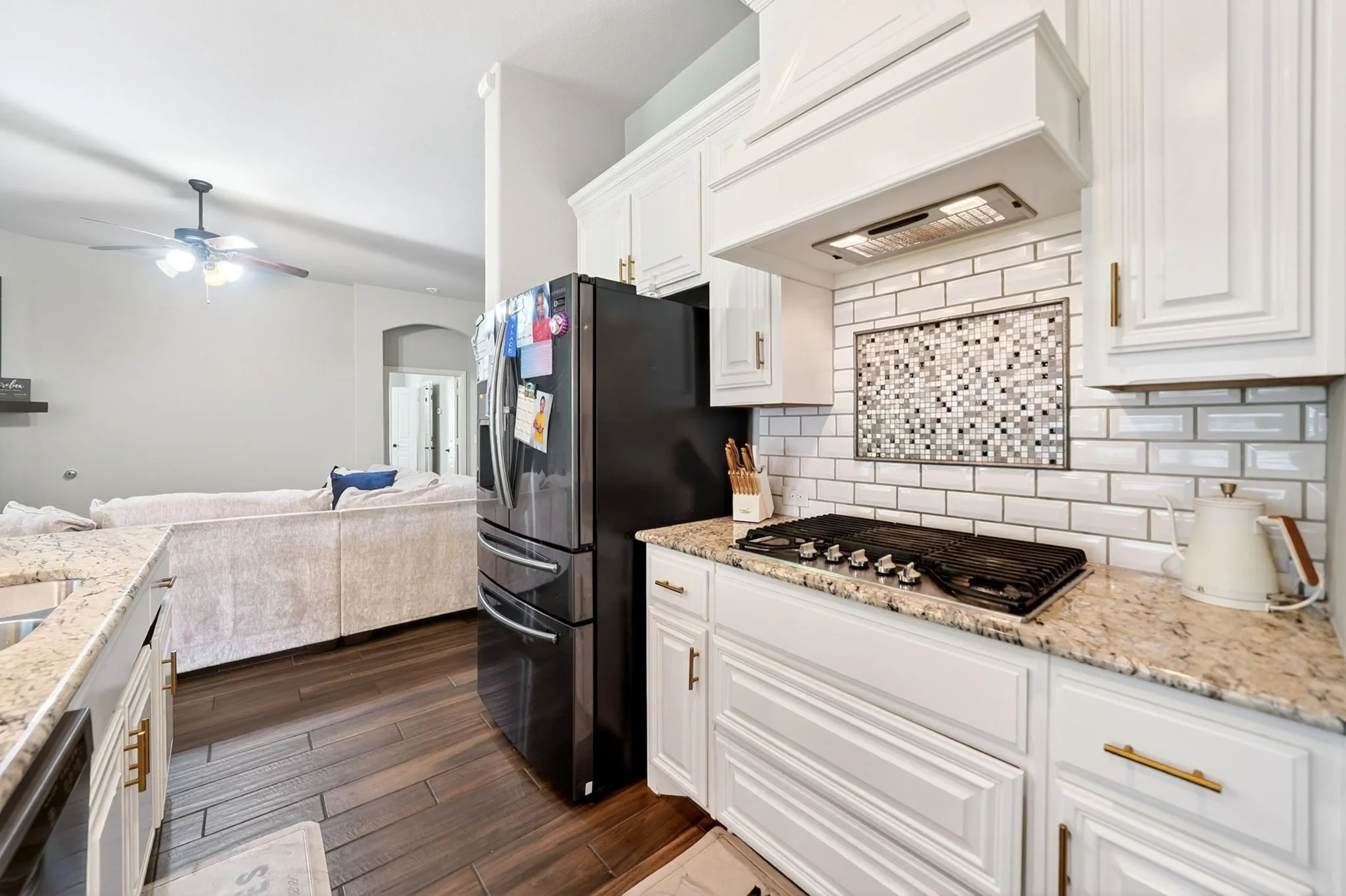 Kitchen featuring light-toned countertops, white subway tile backsplash, white cabinetry with gold-finish hardware, and a built-in cooktop with an overhead range hood