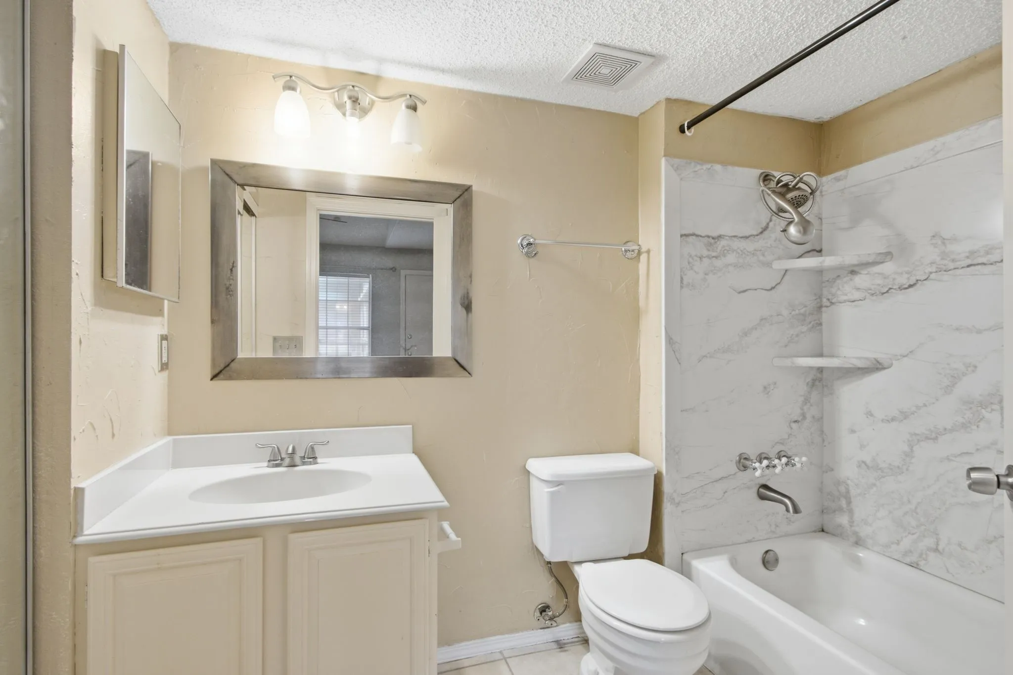 Full bathroom featuring a single vanity with a white countertop, a mirror with a wood-finish frame, a tub/shower combination with a marbled surround, and recessed shelving